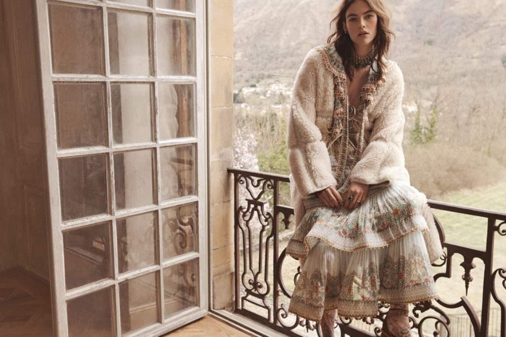 Model sat on balcony wearing a long dress and fluffy jacket