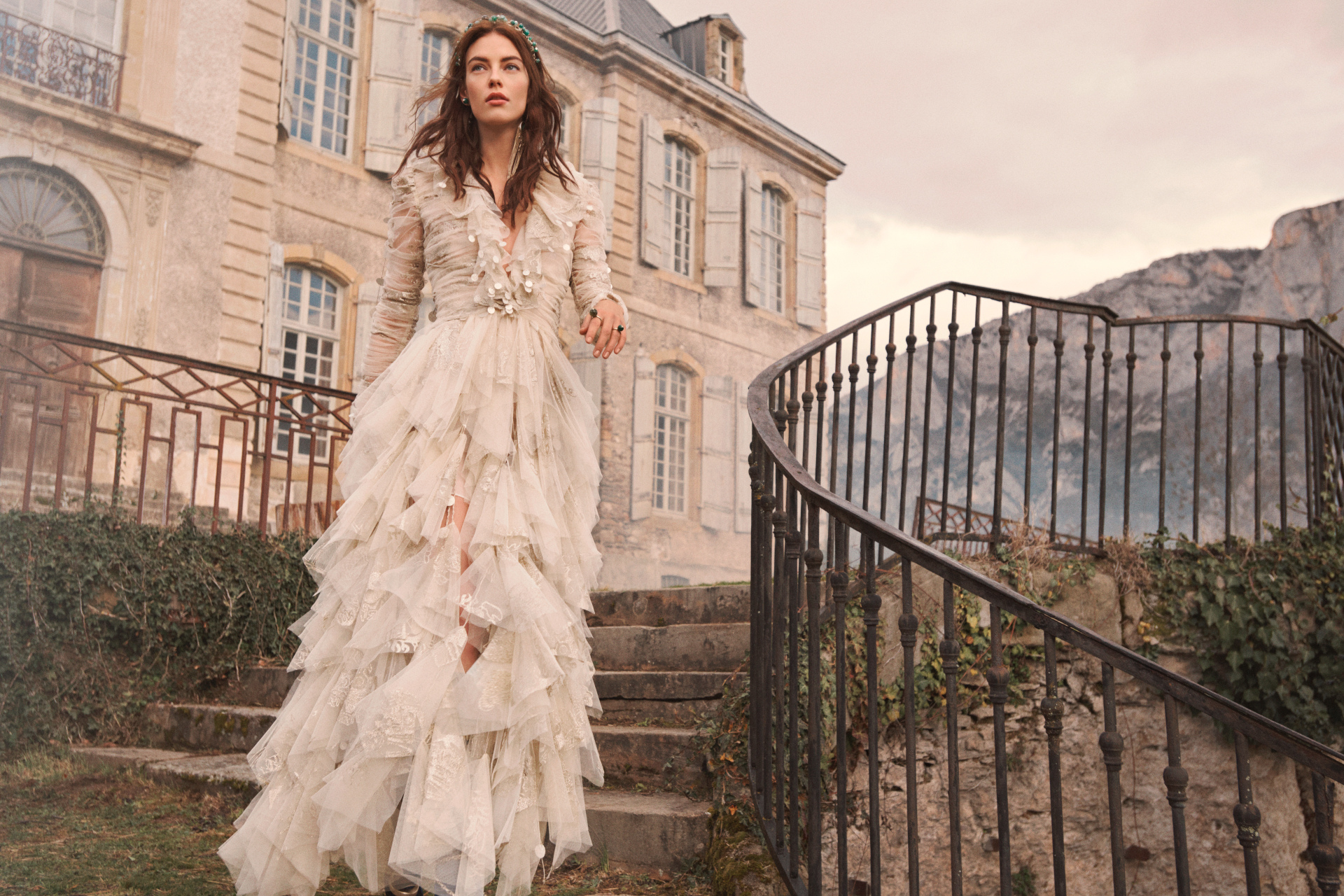 Model walking down front steps with black railings, wearing a long white ruffled gown
