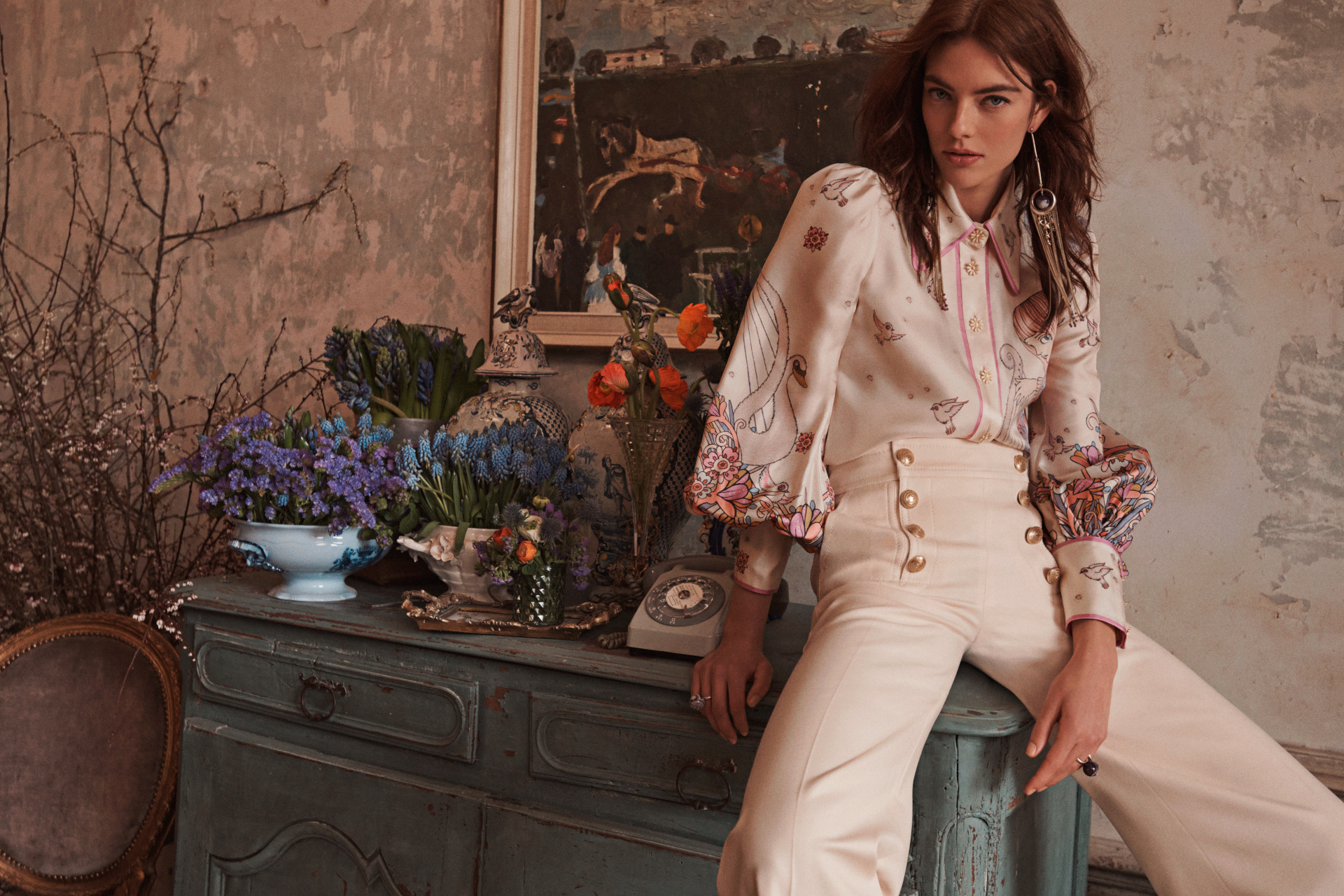 Model sat on blue set of drawers wearing a white puff-sleeve shirt and white trousers