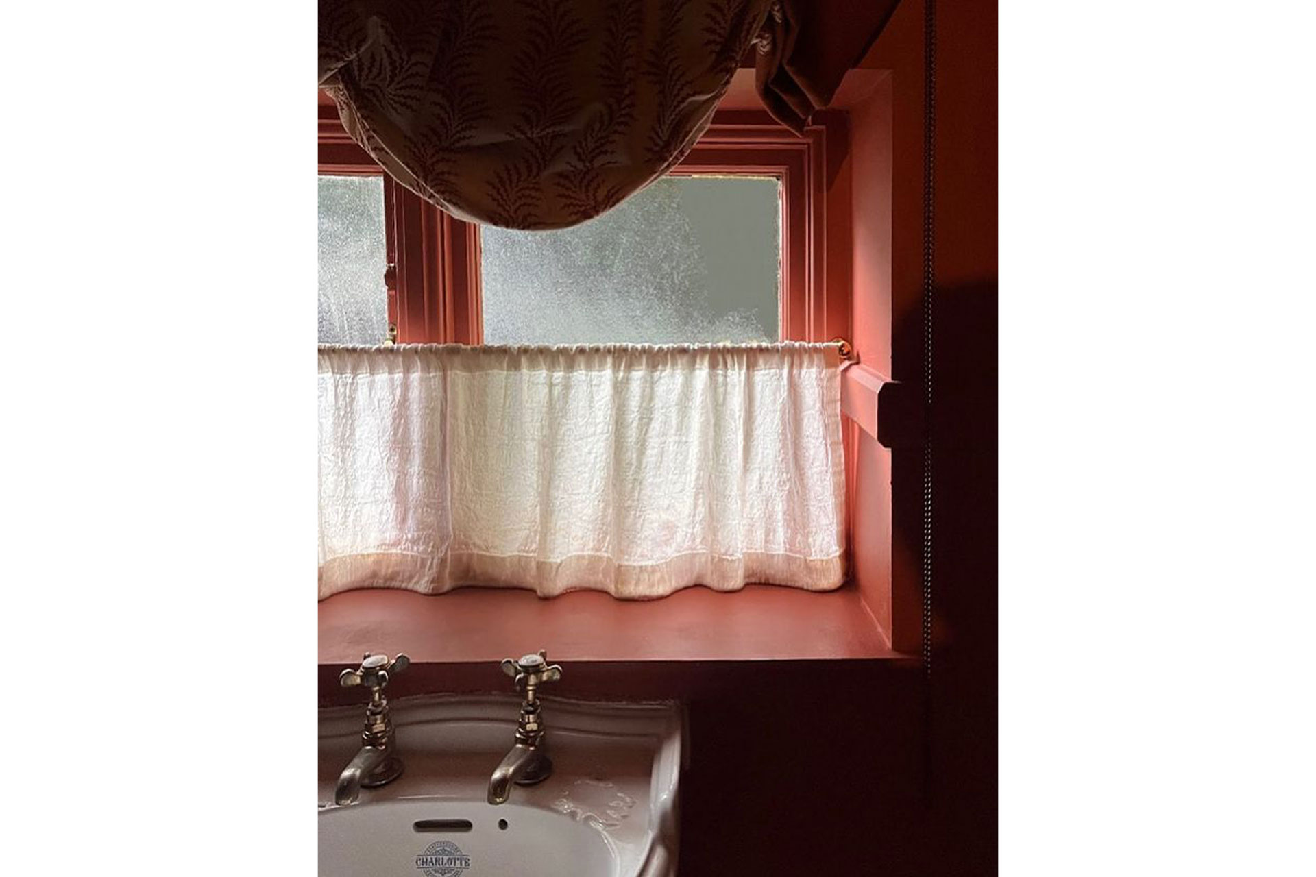 a window of a pink bathroom with a small white curtain next to the sink