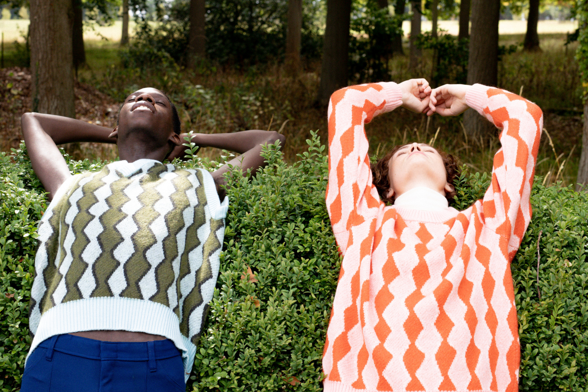 Two people in diamond pttern knitwear laying back on hedgerows