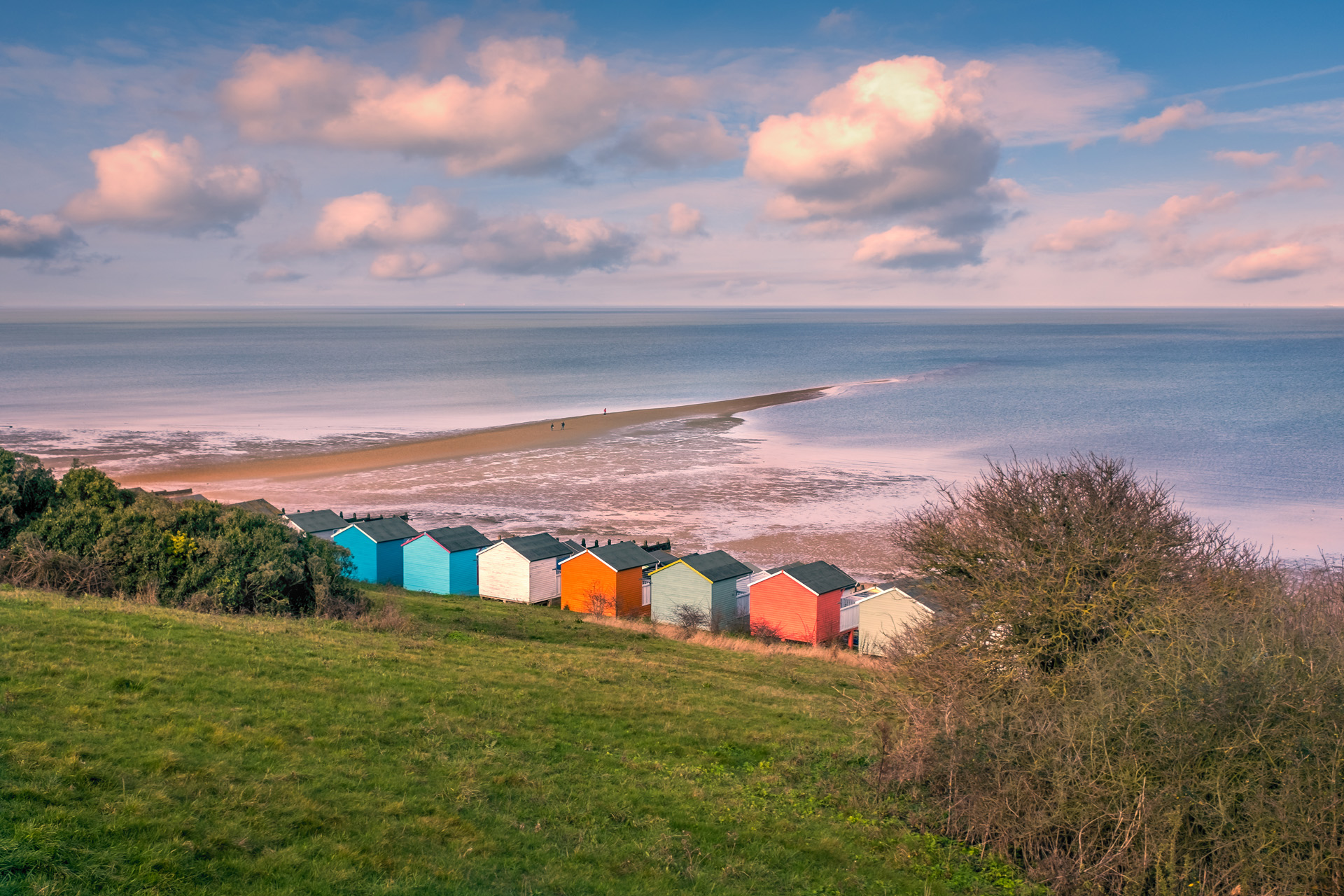 Impressive winter clouds in a cool blue sky over the beach huts and natural spit of land that stretches out to sea on the beach in Tankerton, Whitstable, Kent, UK. A three people are strolling on the natural and locally named 'Street'