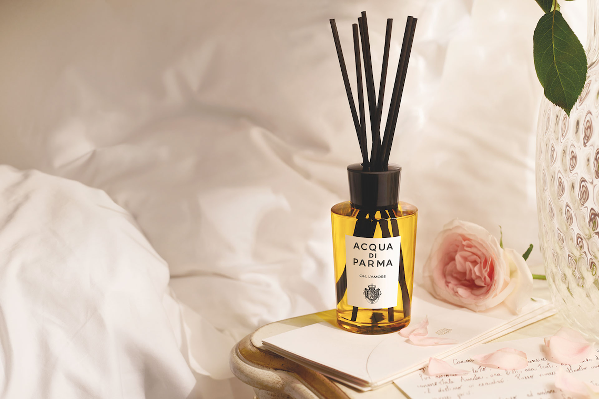 An Aqua Di Parma diffuser next to a bed on a bedside table with a rose on it