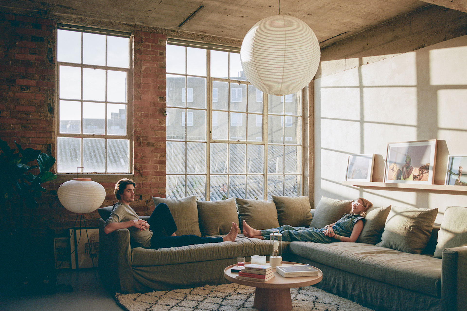 Jack Harries' and Alice Aedy lie on a sage green sofa in the living room of their home in Hackney. The room has floor to ceiling windows, a cocoon ceiling light, a textured checked rug, and a shaft of light is coming through in a refurbished concrete space.