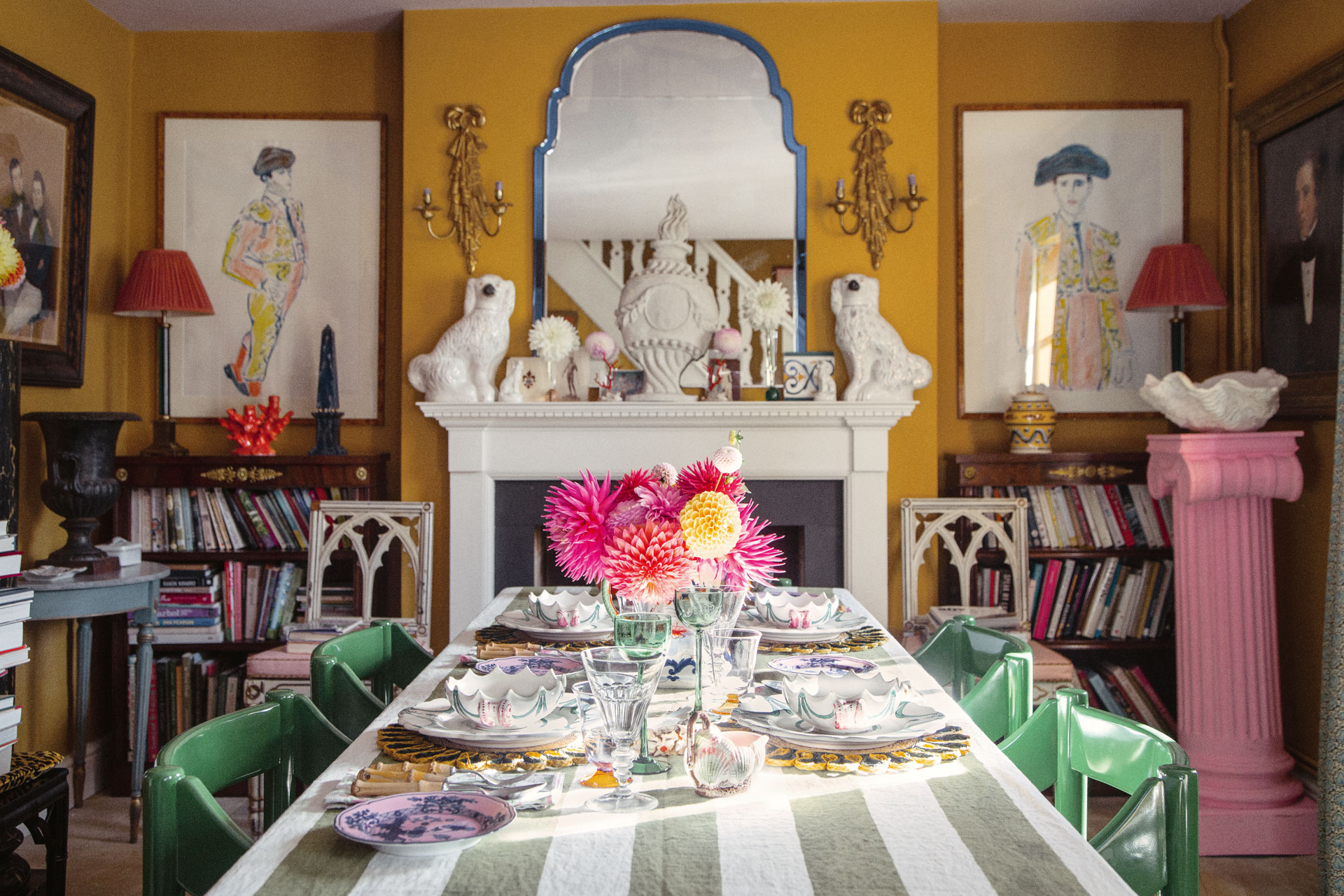 Luke Edward Hall's dining room, featuring a stripy tablecloth, pink pillar, yellow walls, green chairs, a mirror with a blue border that's flanked by two porcelain cocker spaniels