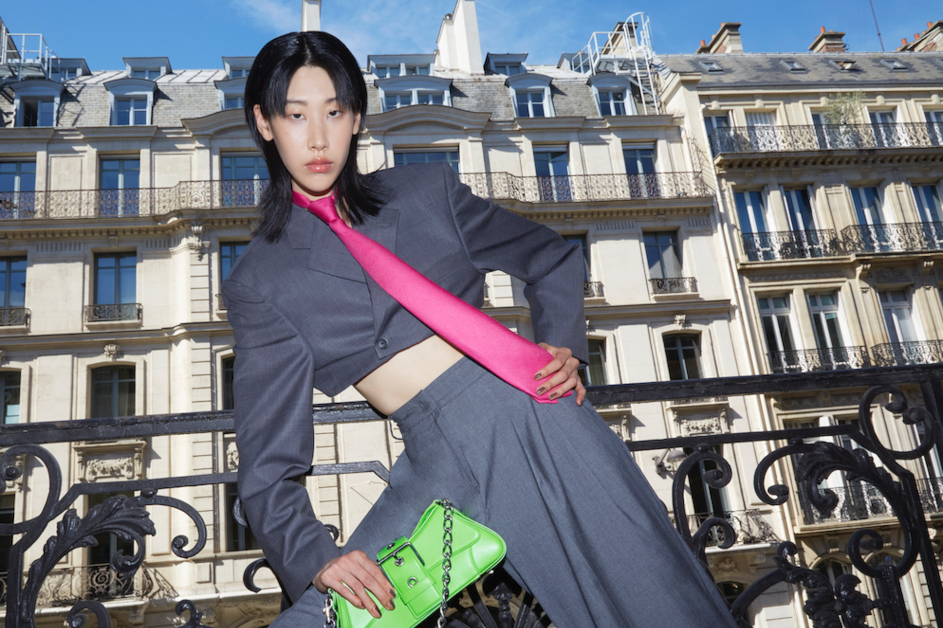 Model in grey cropped suit and pink tie with green handbag leaning on one leg, with Parisian architecture in the background