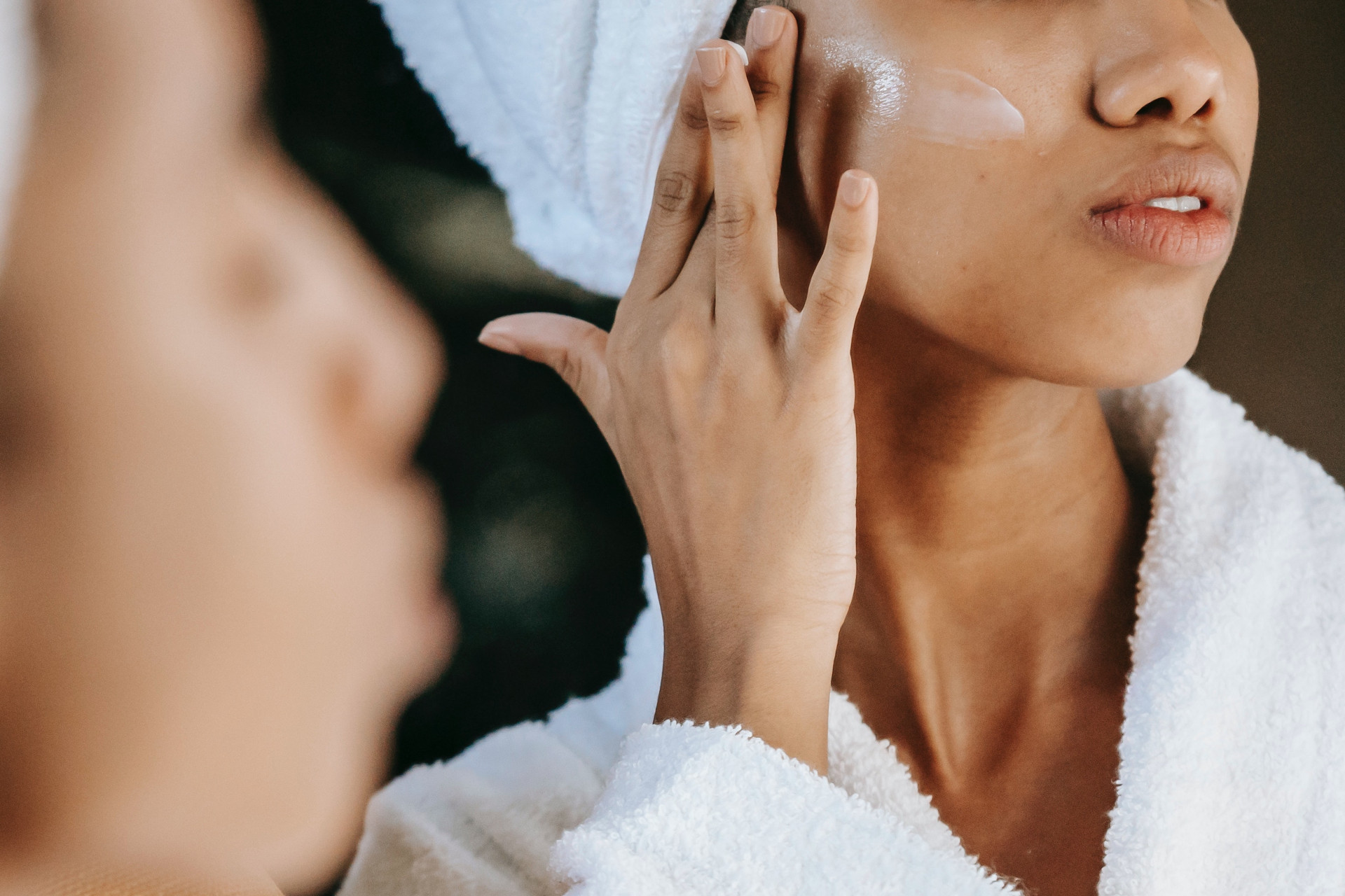 Woman rubbing cream on face while wearing hair towel and white robe