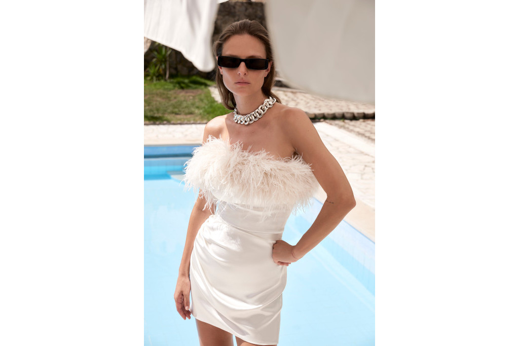 Close up of model in white feather mini dress and sunglasses, stood in front of pool