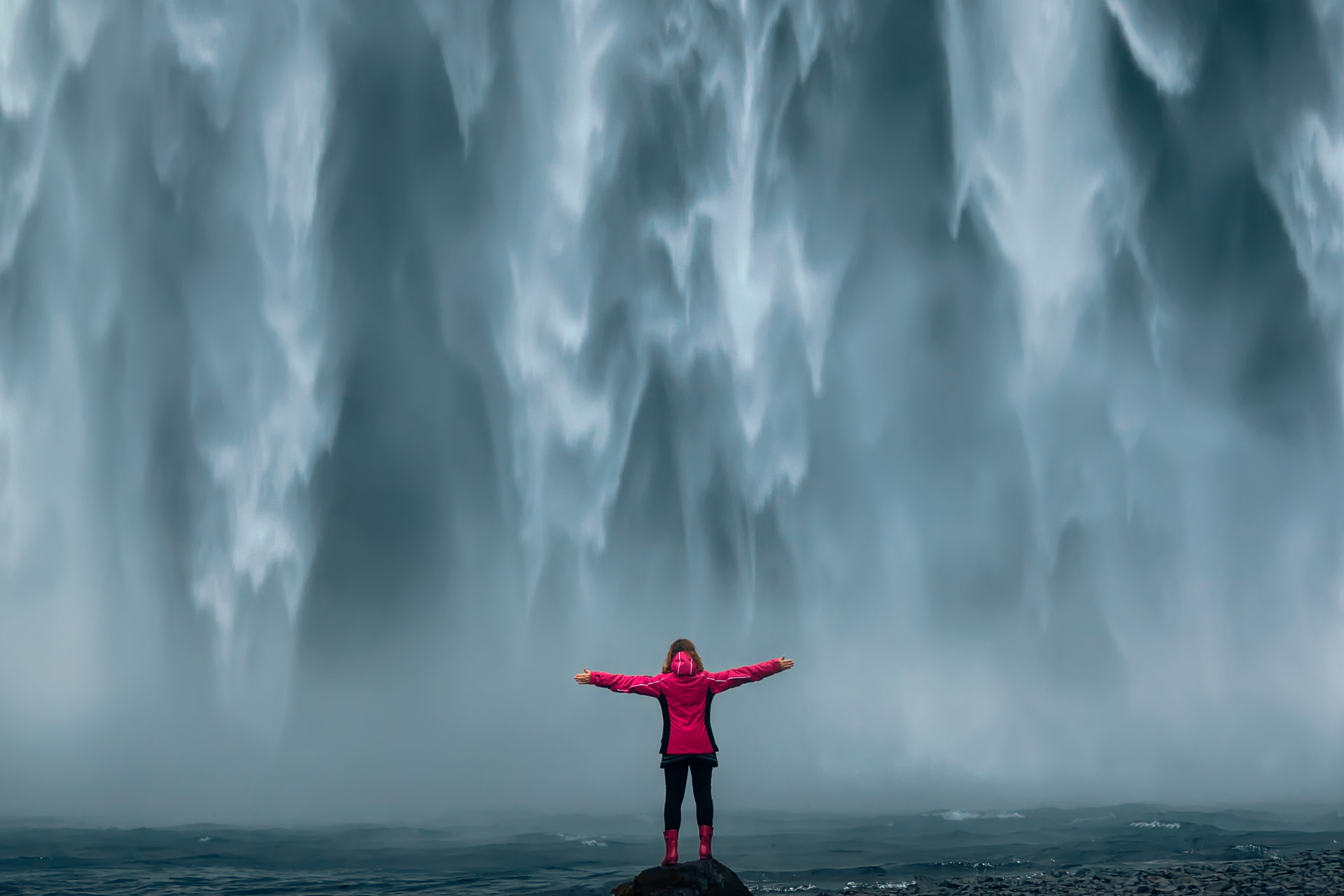 Iceland landscape photo of brave girl who proudly standing with his arms raised in front of water wall of mighty waterfall.