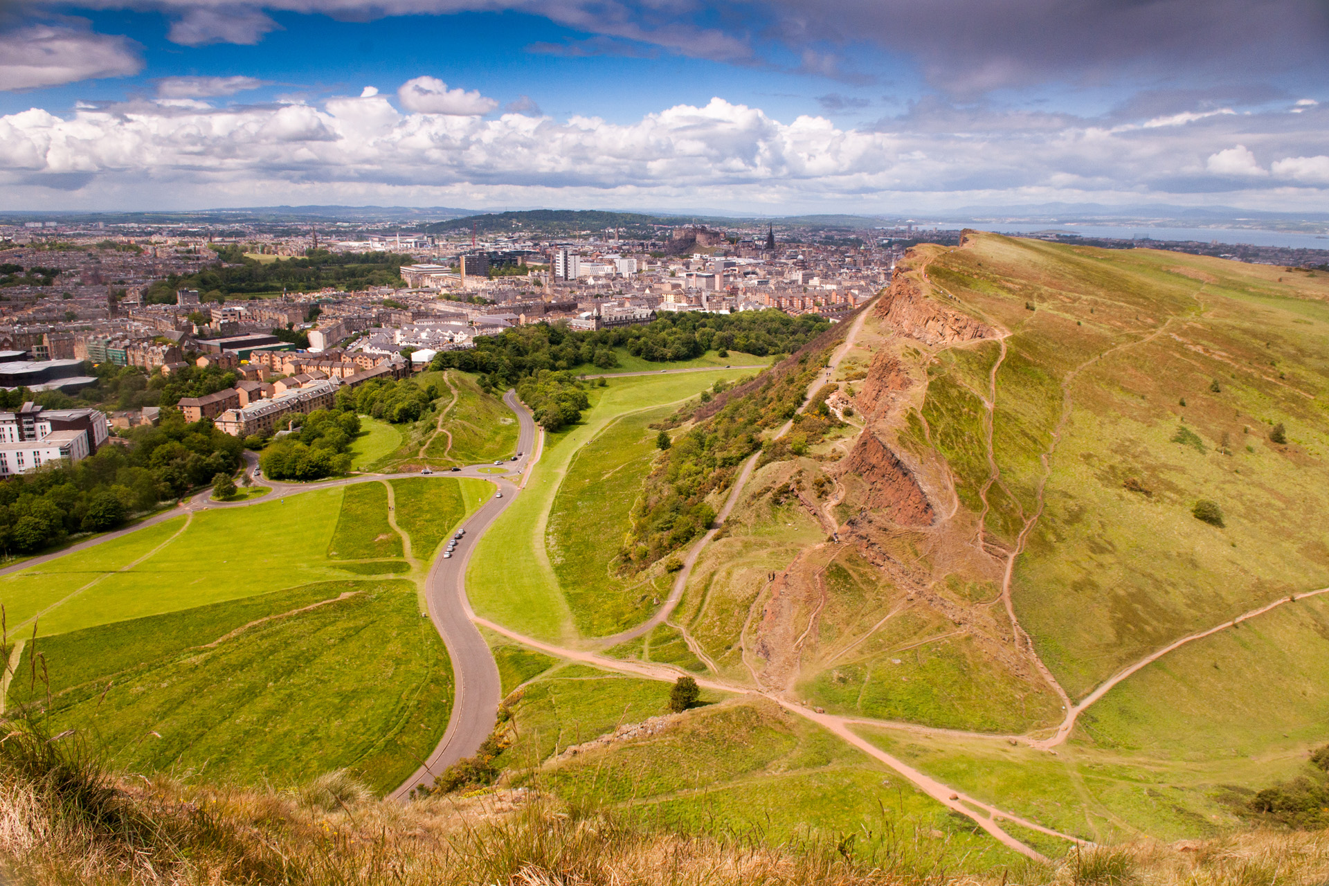 The Edinburgh cityscape, including the Old Town and Edinburgh Castle, seen from Arthur's Seat on a sunny summer's day, with Salisbury Crags and Holyrood Park in the foreground.