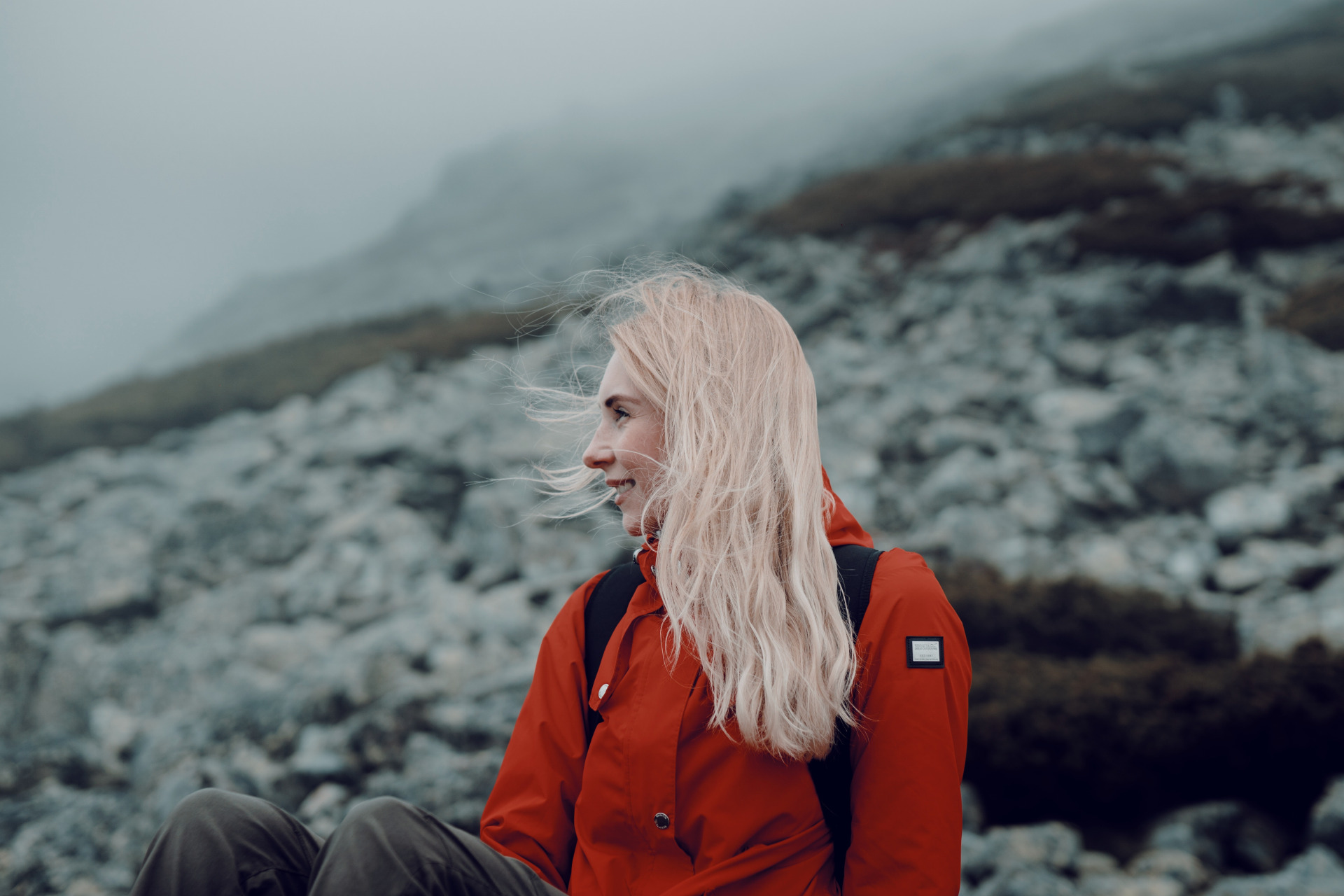 Woman in red jacket with blonde hair sat on rocky hill - hair trends 2023