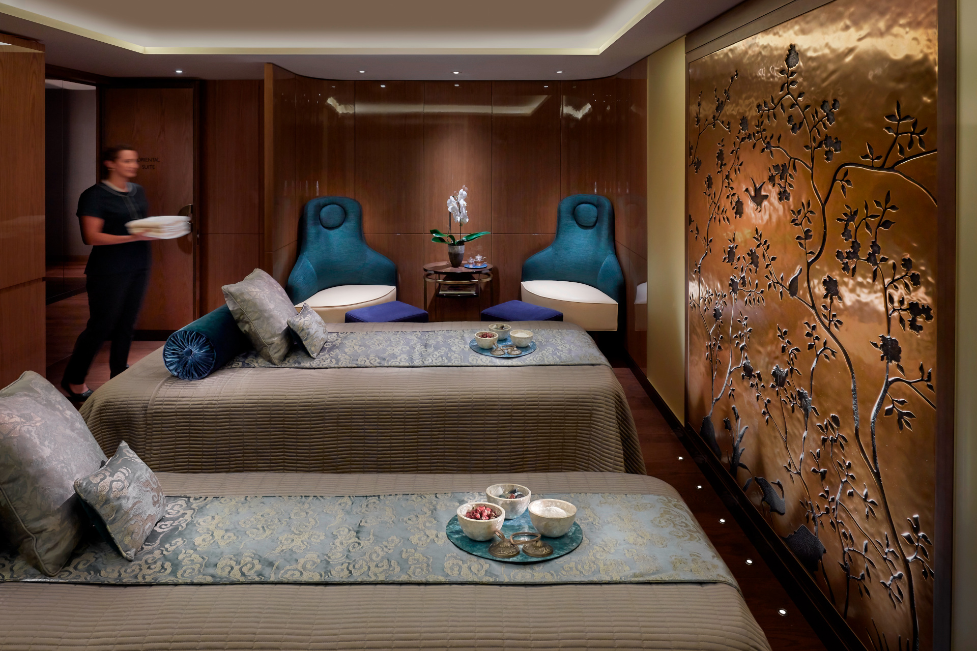 Spa treatment room with two beds, blue chairs and gold walls, with attendant walking in with towels