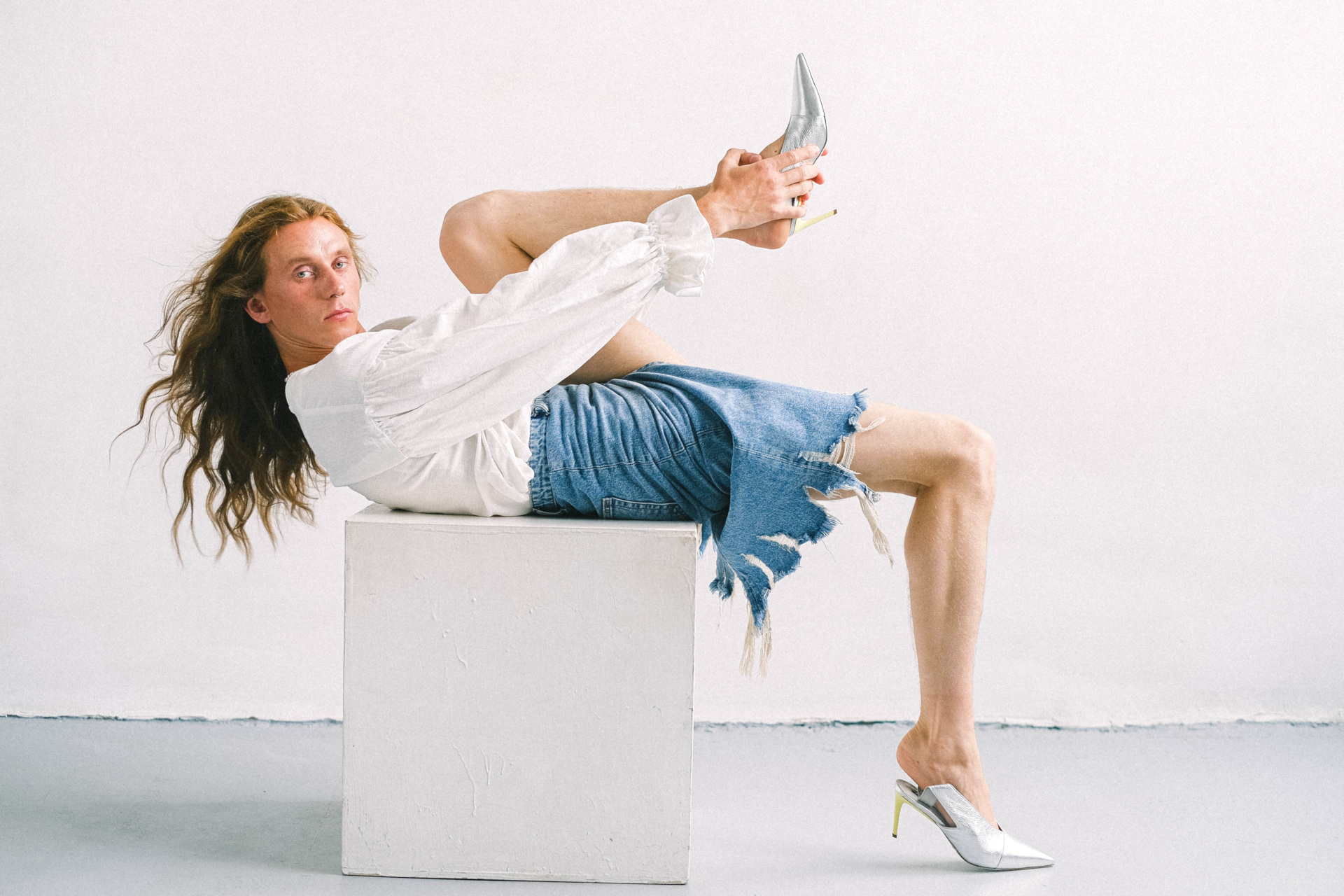 Genderfluid person leaning back on white block wearing shirt and denim skirt