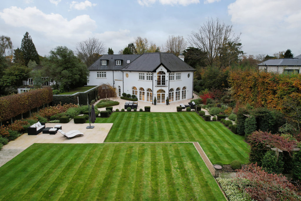 Manicured lawns with a patio seating area and a large white house with curved windows and grey roof.