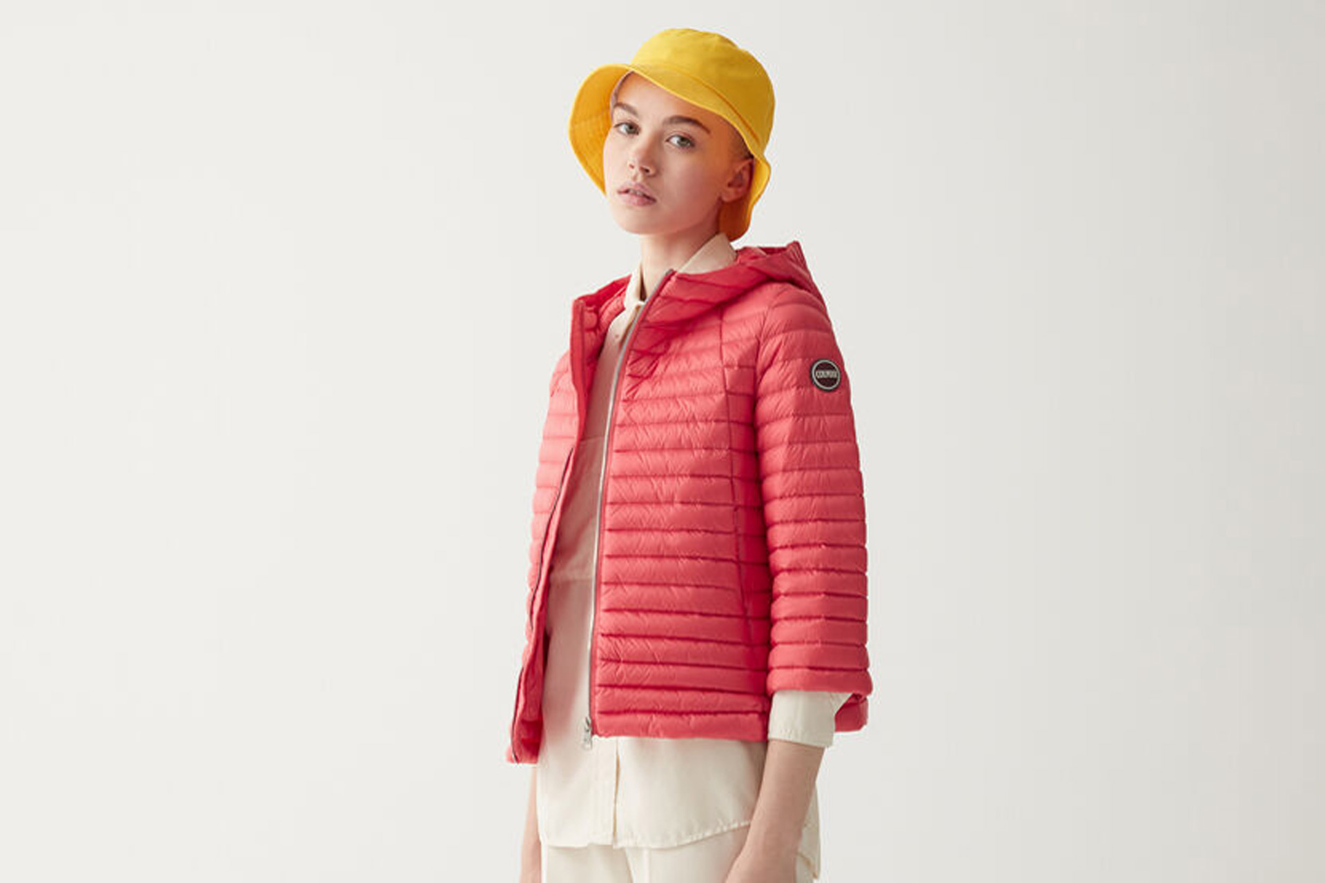 Salmon pink padded down cape jacket worn with white trousers and a yellow bucket hat.