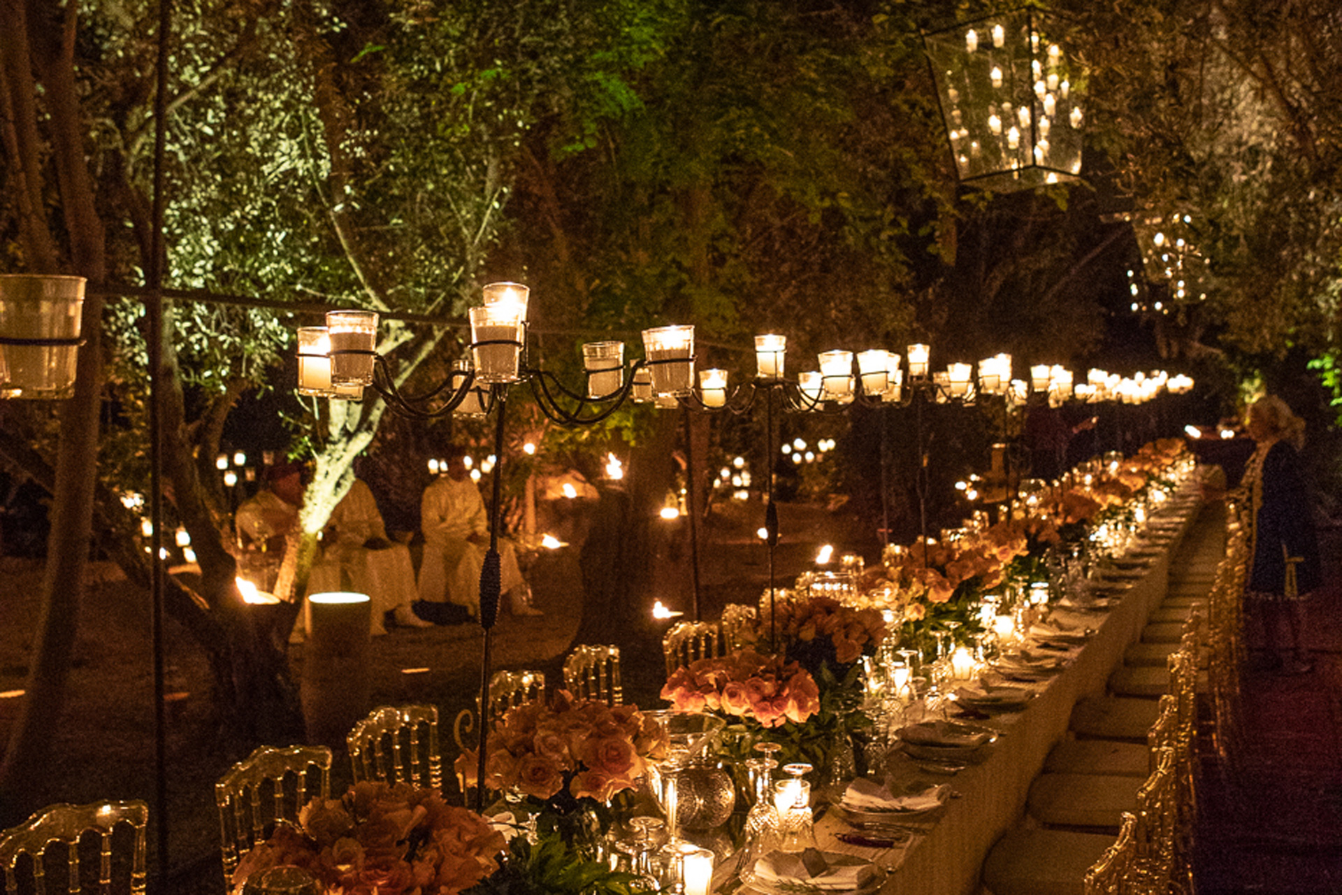 Long candlelit table outside surrounded by greenery, with lights hanging above
