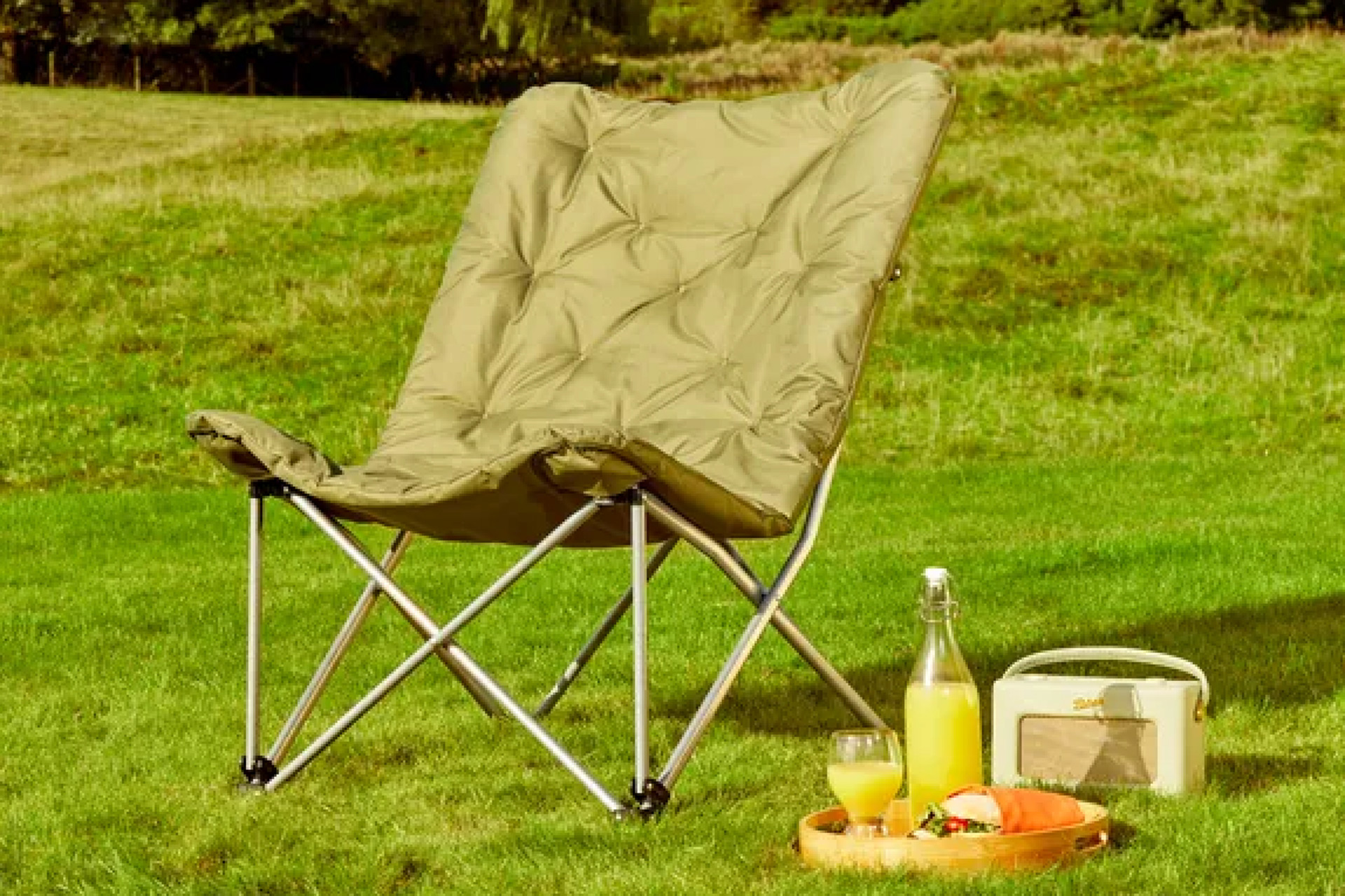 Olive green camping chair in field with radio, juice bottle and cup beside it