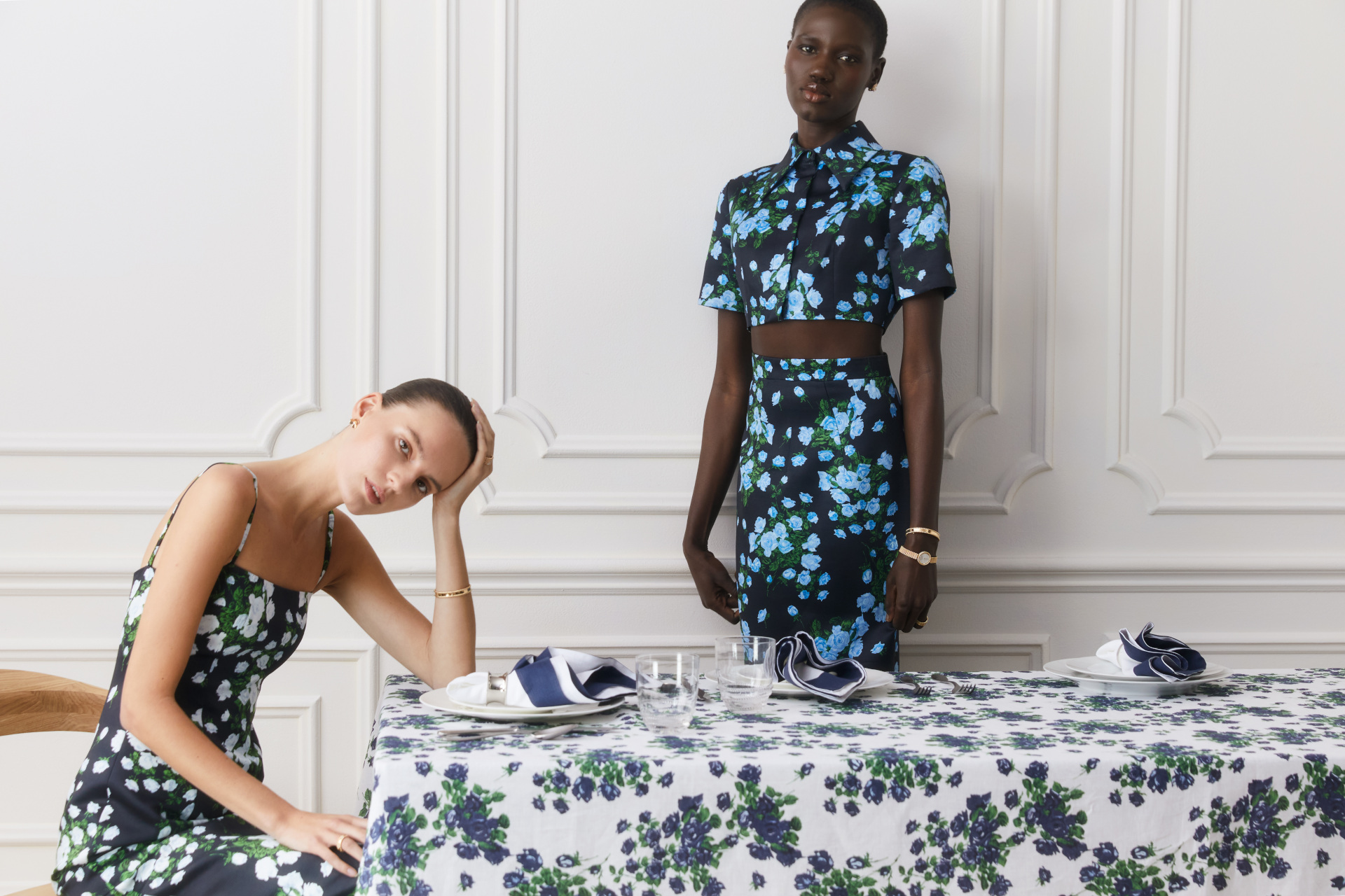 Two women in floral clothing sat and stood around table laid with floral tablecloth