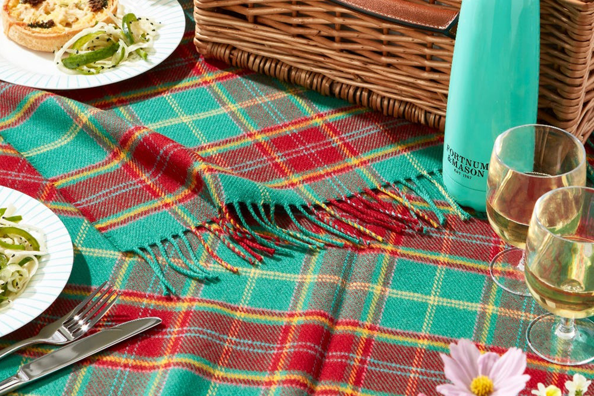 Sea green and red tartan picnic blanket with plates of food, picnic hamper, and Fortnum & Mason water bottle on top