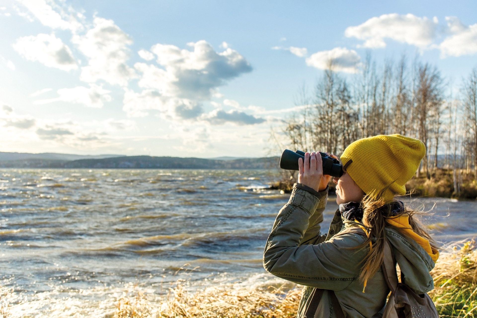 Woman with yellow beanie looking through binoculars onto a lake