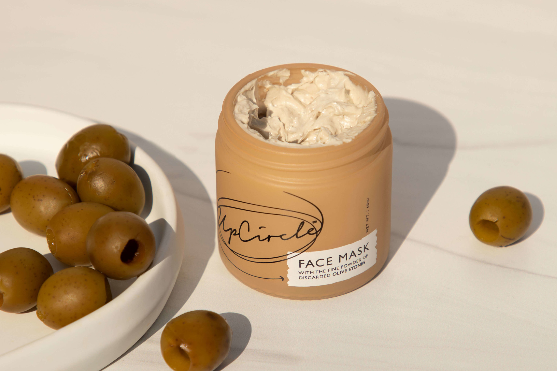 Light brown tub of face cream next to plate of olives