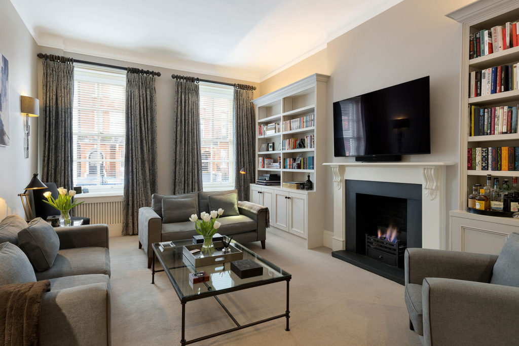 Modern living room with two large windows and grey furniture. Fireplace below television and two bookshelves either side.