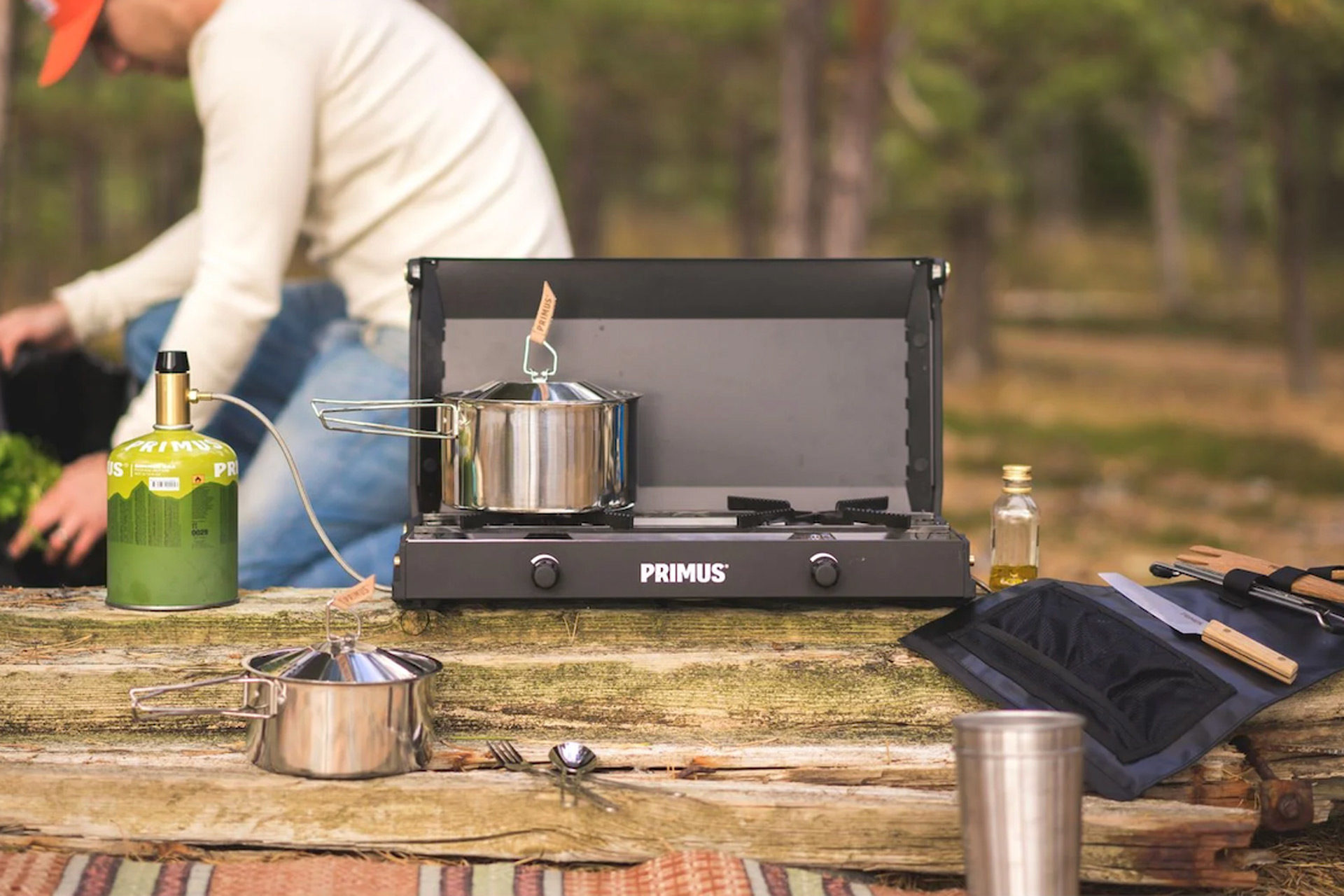 Camping stove with pots, cutlery and gas beside it, with a man in orange cap sitting in the background