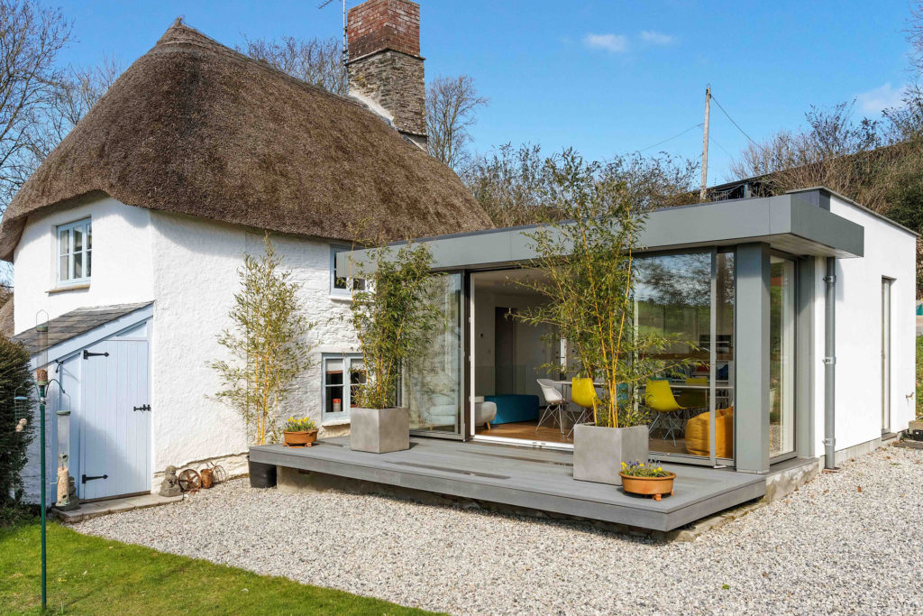 White thatched cottage with modern extension featuring large glass doors and a grey terrace.