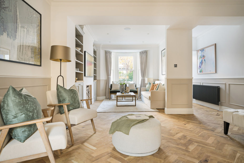Large living room with parquet flooring, white boucle armchairs and footstools, and a bay window with taupe curtains.
