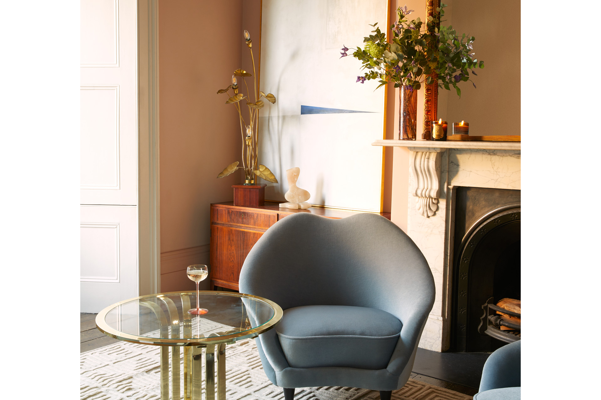 Living room with dusty blue chair, glass side table and mantlepiece with vase of flowers