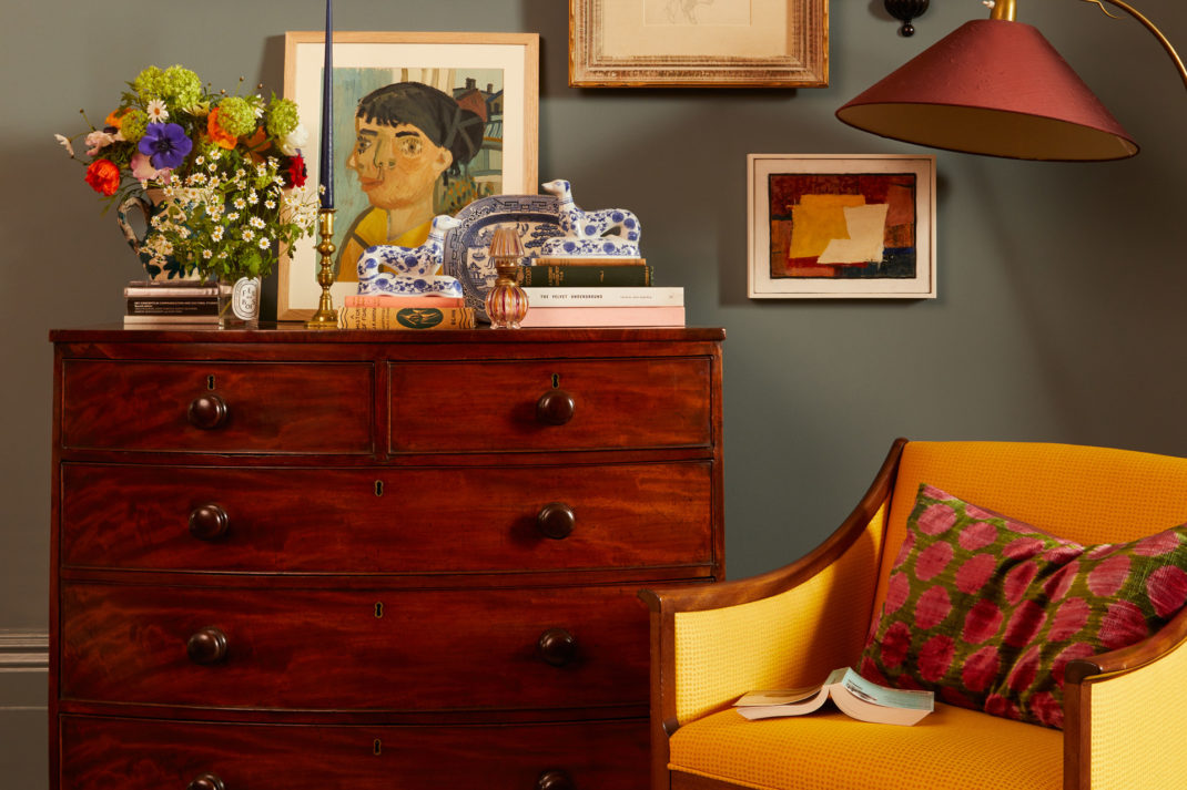 Blue living room with mahogany chst of drawers, bright yellow armchair, pink lamp, and colourful wall art