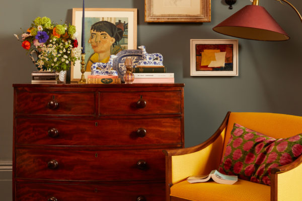 Blue living room with mahogany chst of drawers, bright yellow armchair, pink lamp, and colourful wall art
