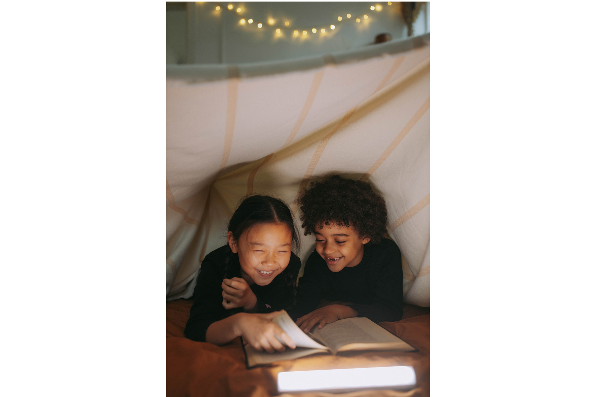 two kids reading a book under a blanket