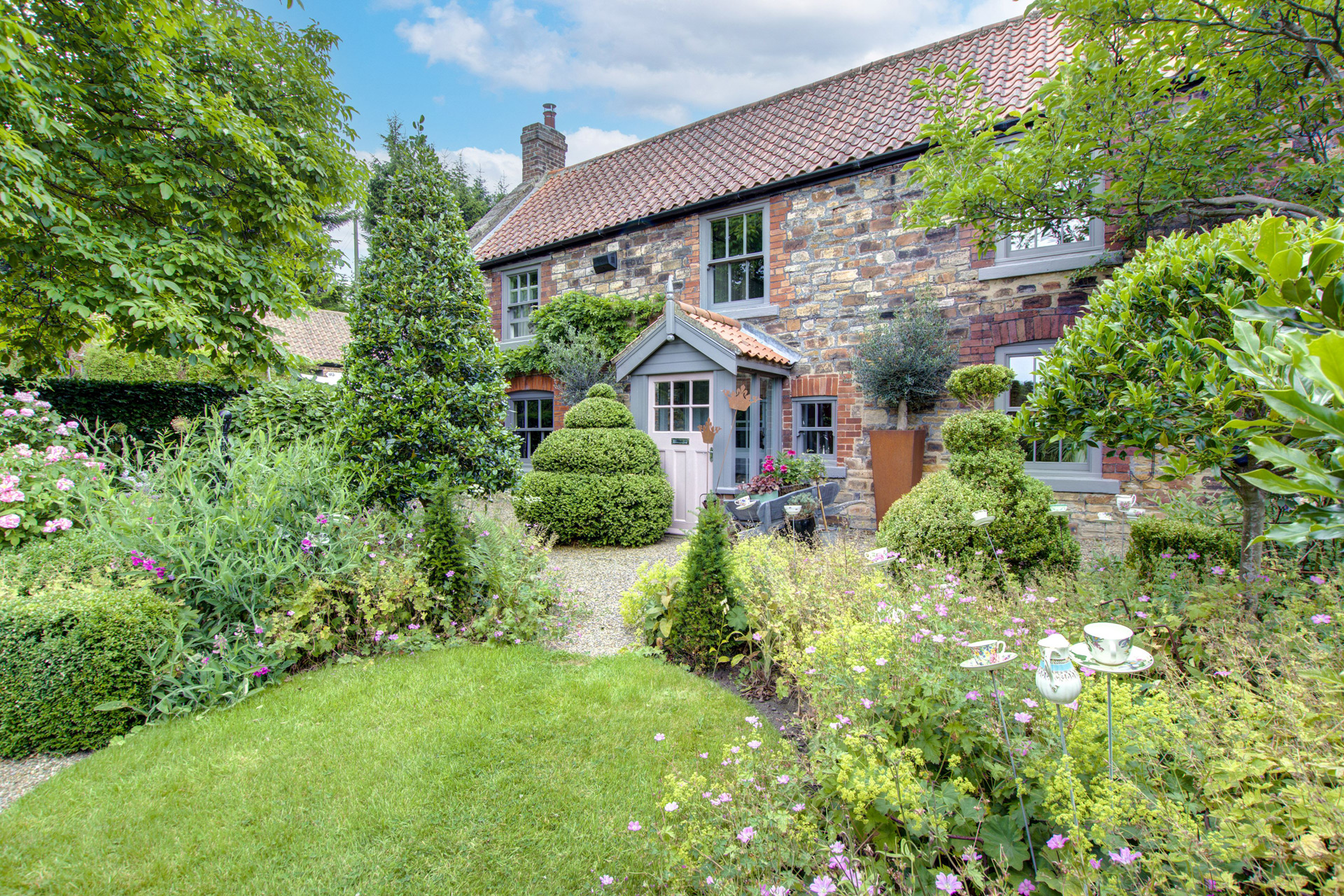 Cobbled stone farmhouse with hedges and wildflowers growing in front.