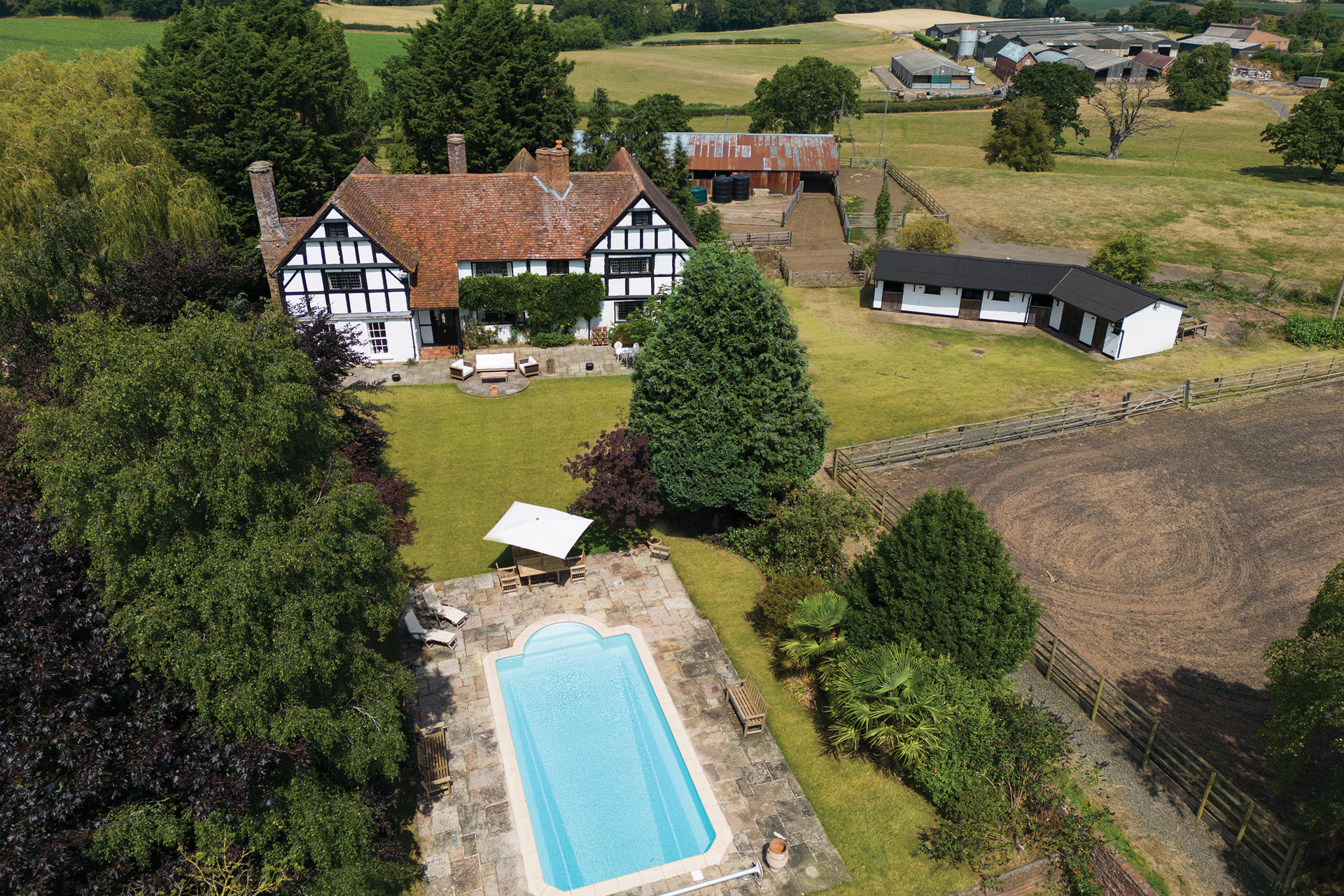 Aerial view of tudor-style farmhouse with fields surrounding and an outdoor swimming pool.