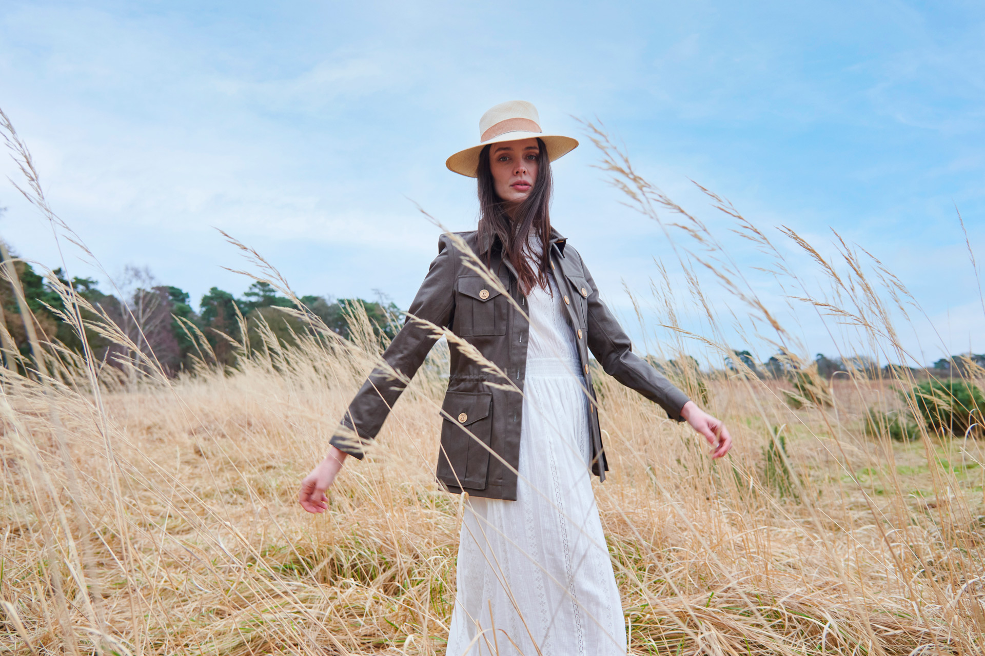 Model in barley field wearing a long white dress, straw hat, and a khaki wax jacket from TROY London/