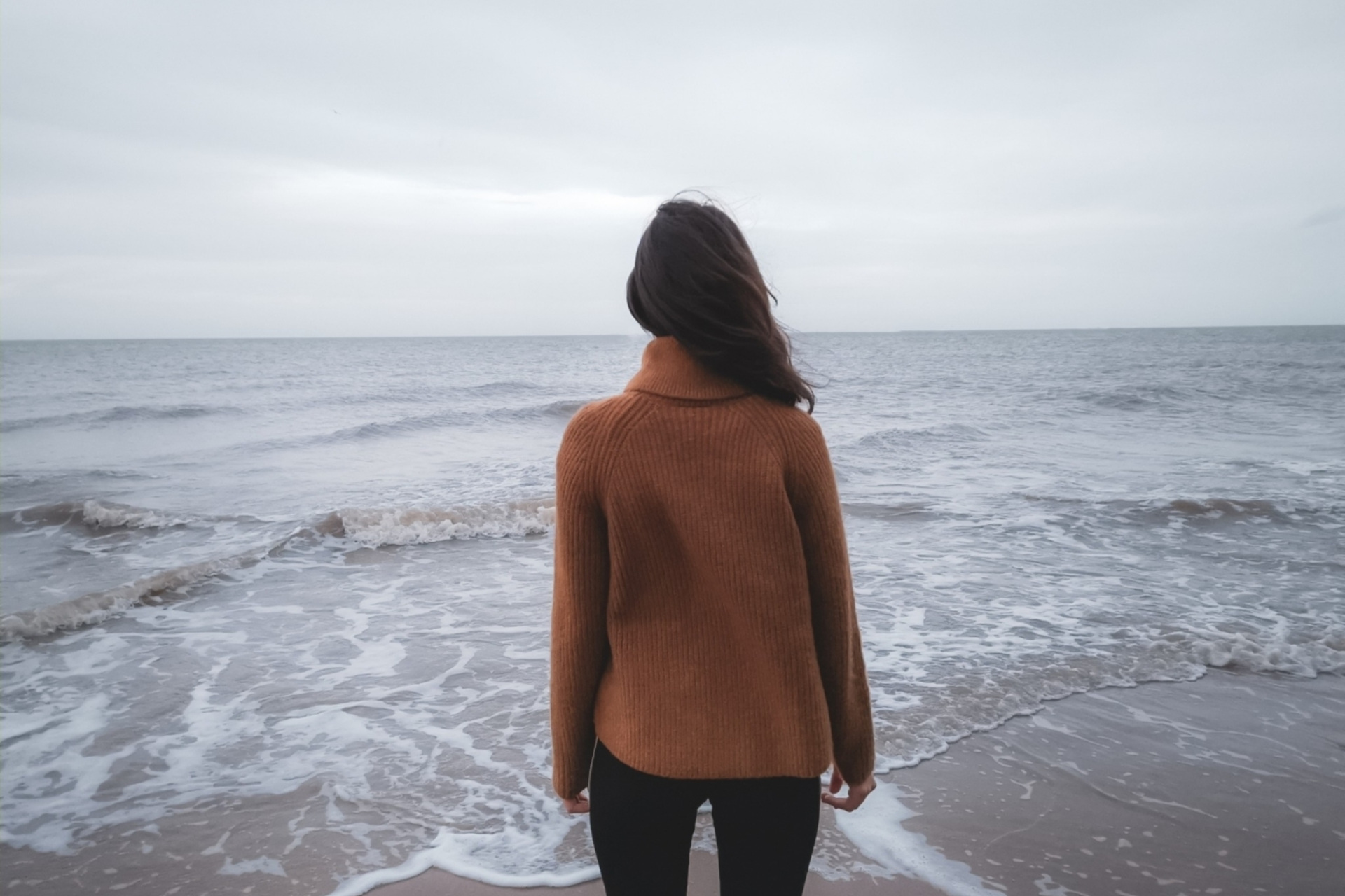 Woman in orange jumper looking out at grey ocean