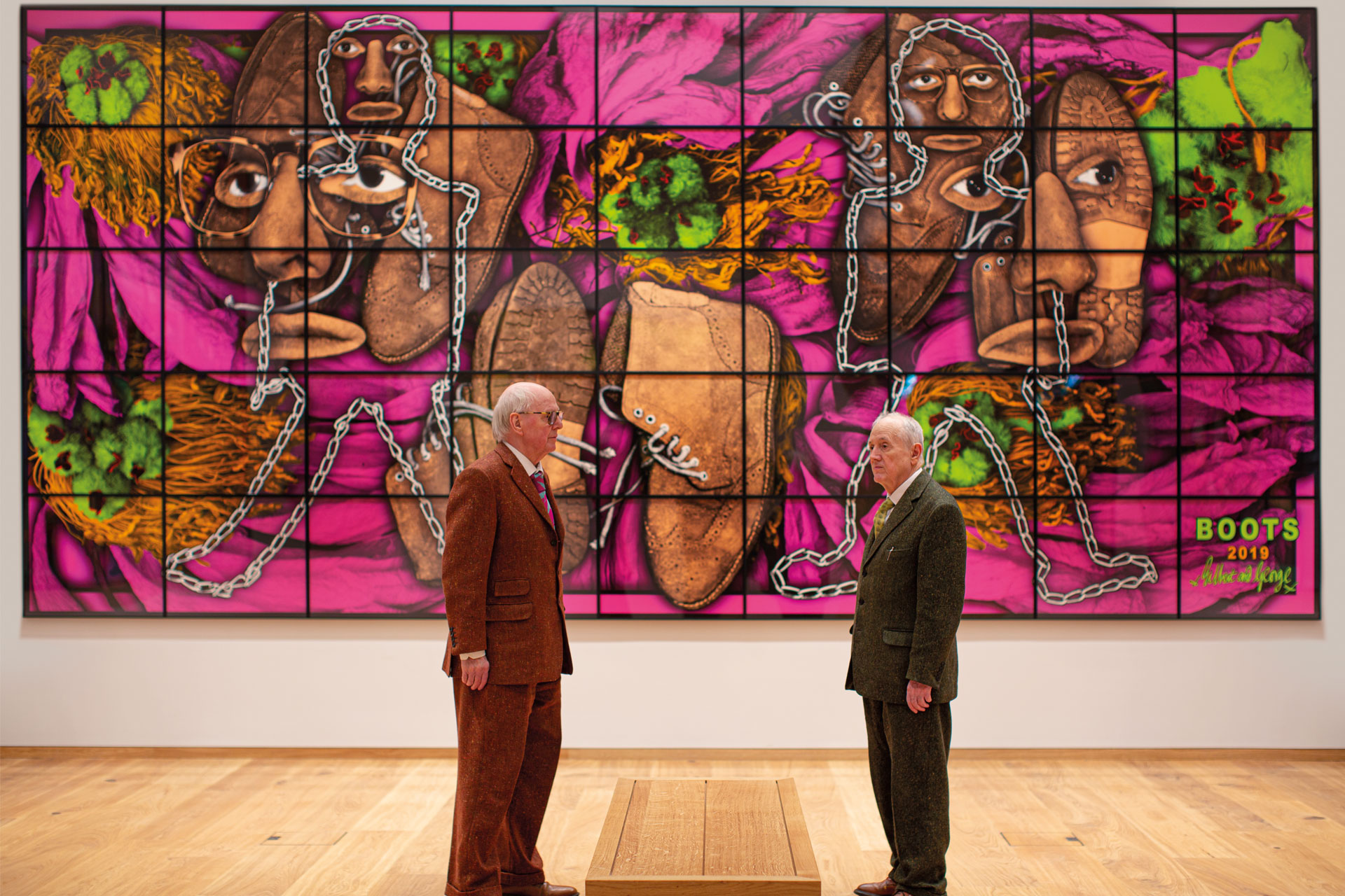Artists Gilbert and George stand in front of some of their work, which is bright, purple and depicts hidden faces constructed from chains
