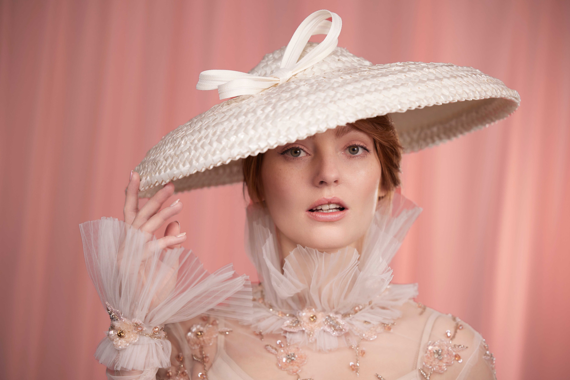 Woman in cream hat and sheer dress in front of pink background