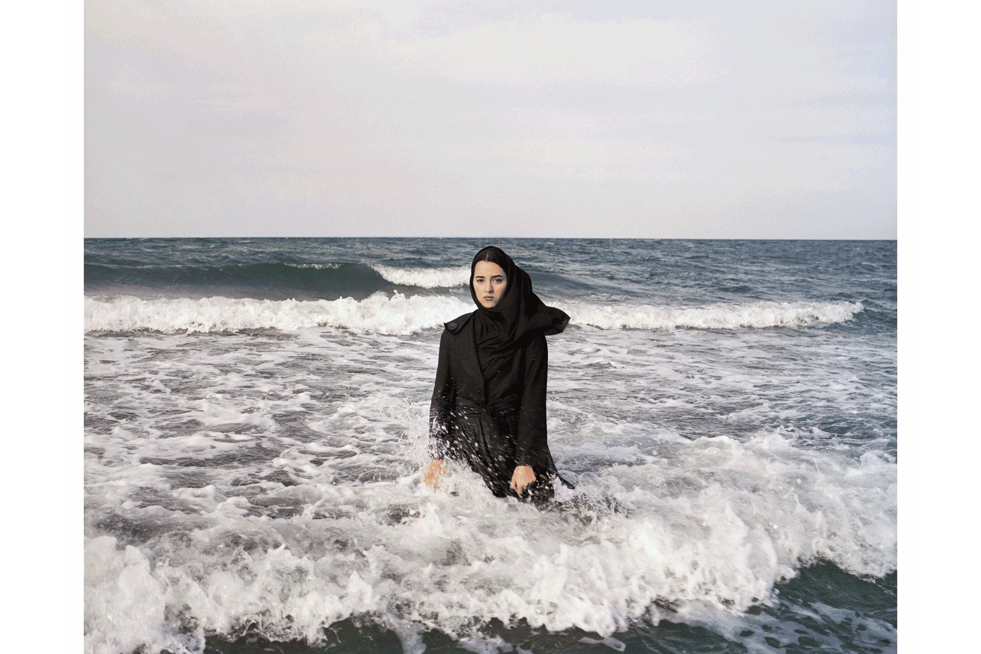 Woman stands in the sea, staring directly at the camera, with her legs submerged and waves behind her.