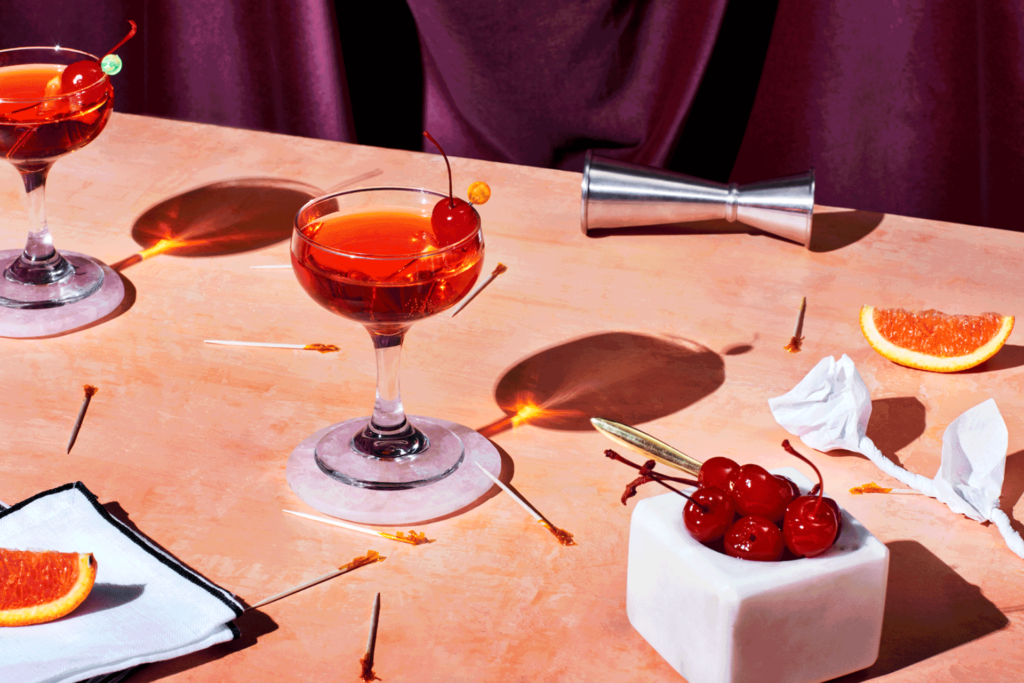 Cherry martinis and slices of orange on a table in front of a purple velvet curtain.