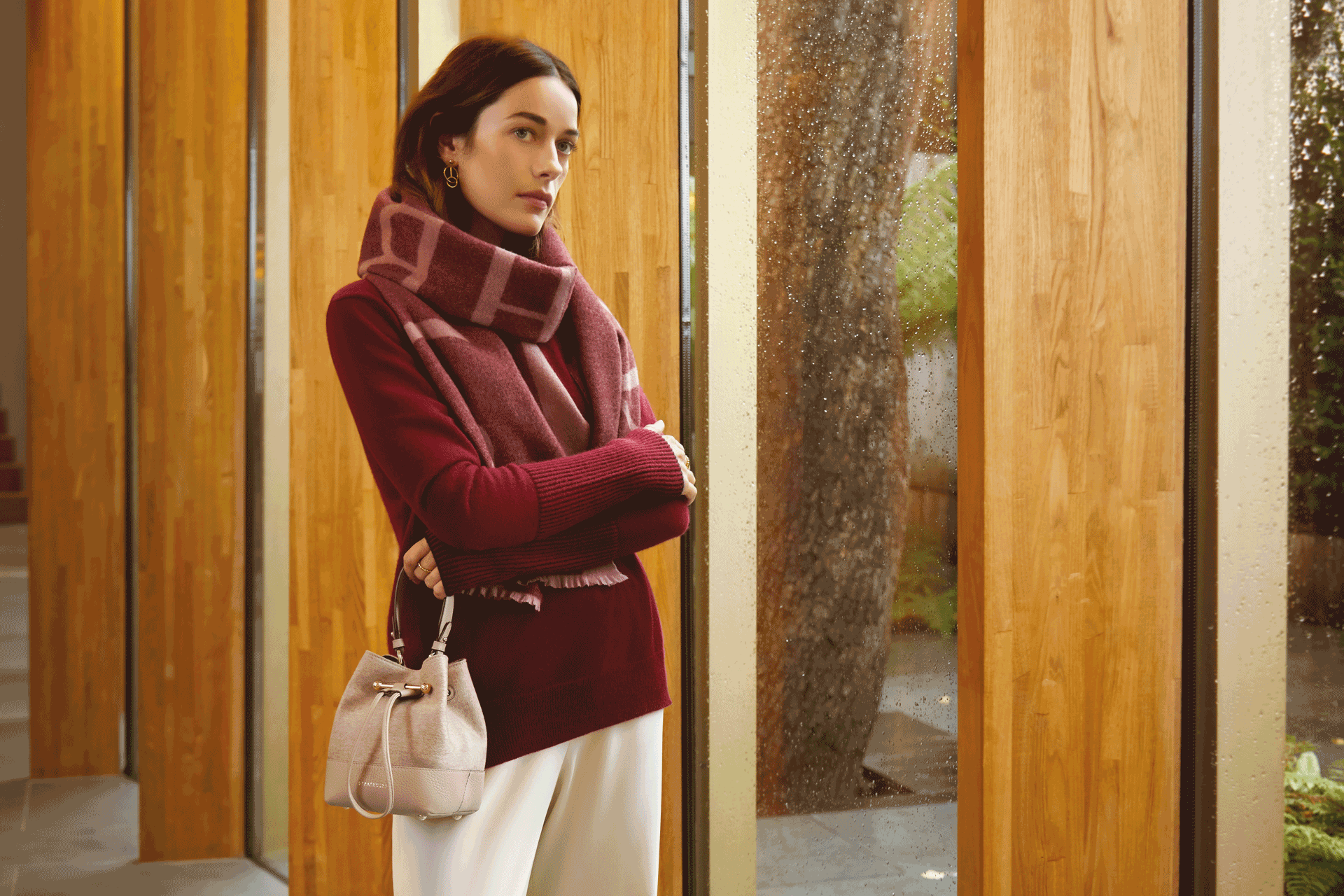 Model wearing a burgundy jumper, striped burgundy Strathberry scarf, a Strathberry bag and Strathberry hoop earrings.