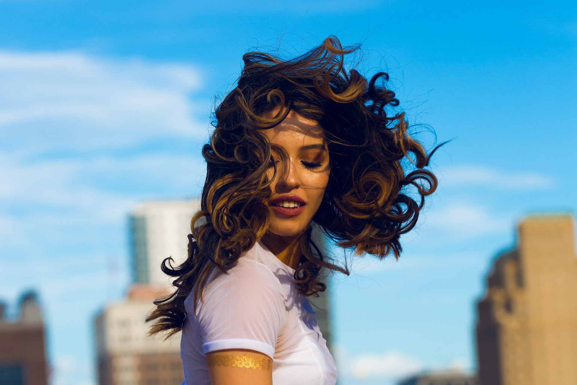 Close up of woman with curly hair being blown by the wind, in front of blue sky background