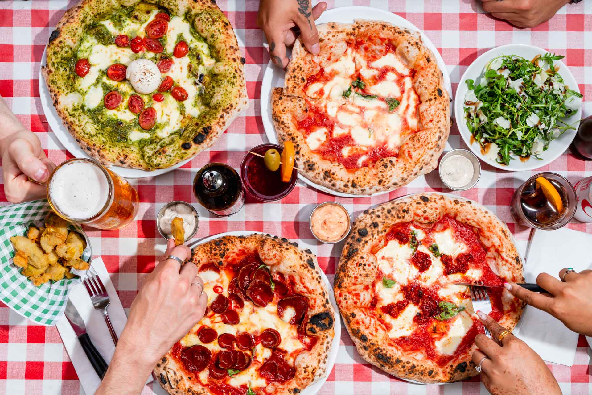 Aerial view of margarita and pepperoni pizzas on a red and white gingham tablecloth.