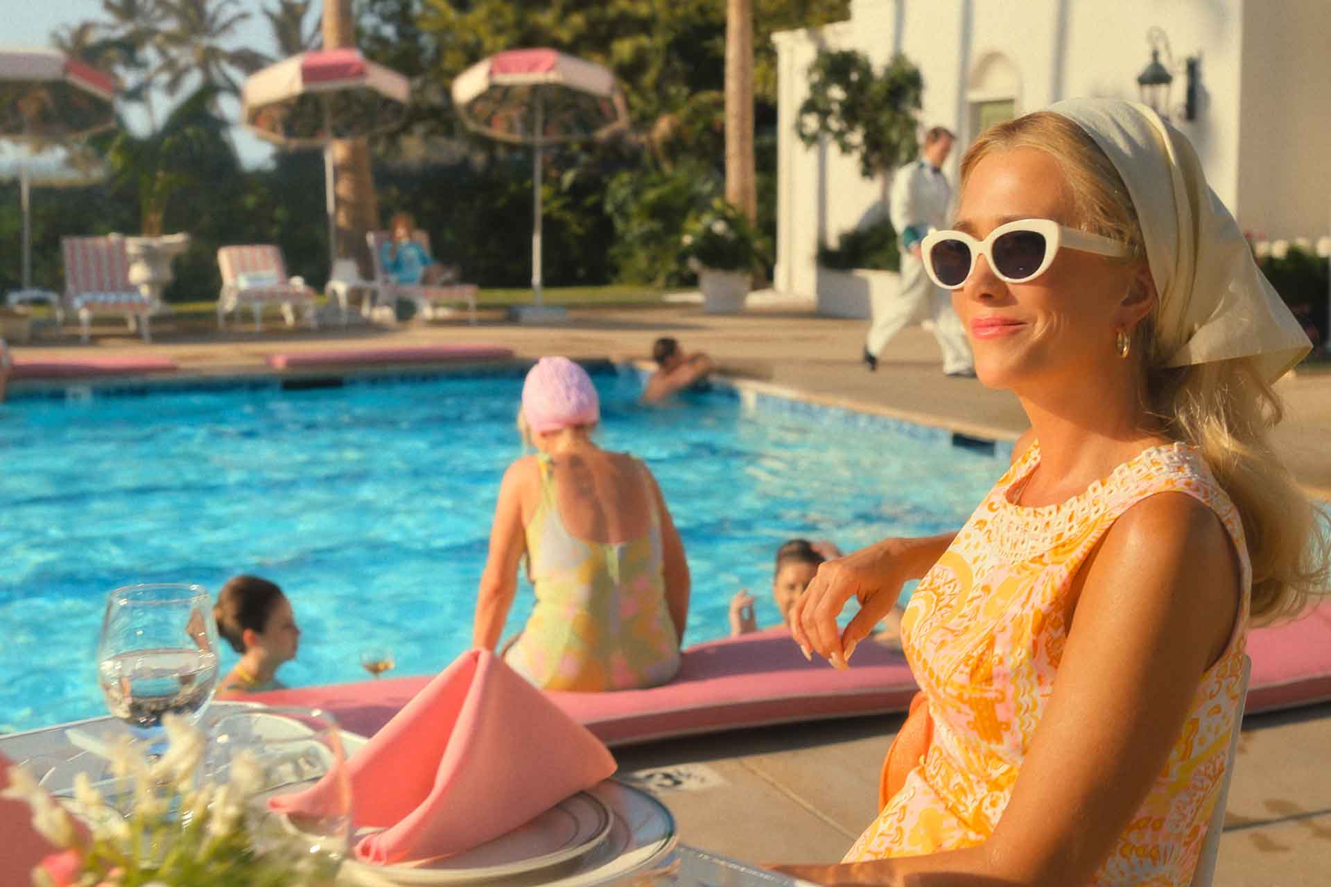 Kristen Wiig sitting beside a swimming pool wearing an orange 1960s dress and headscarf