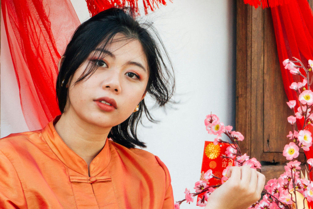 Woman in orange clothing looking forward with red Chinese New Year decorations behind her
