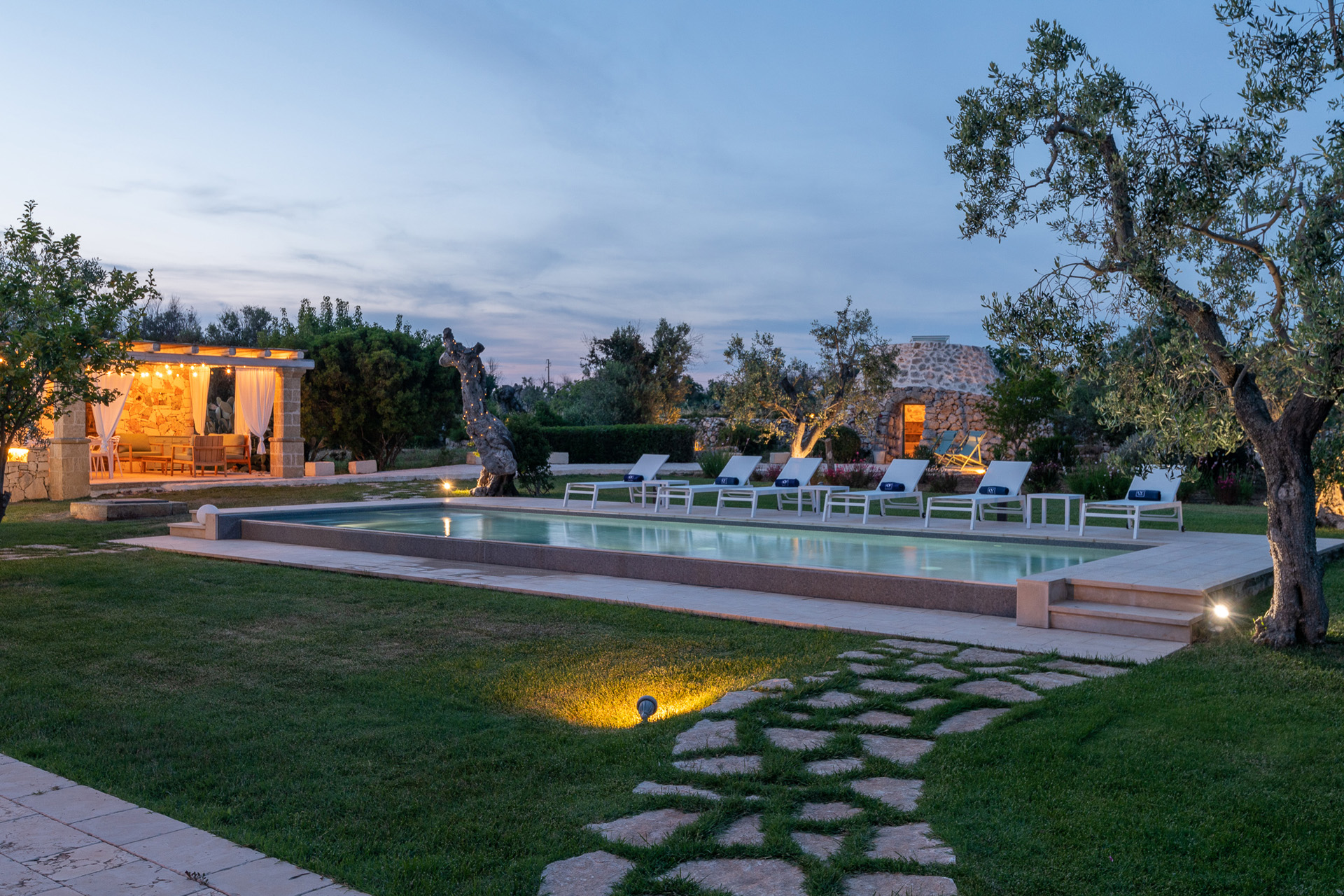 Stone paths leading to a swimming pool flanked by chairs at dusk