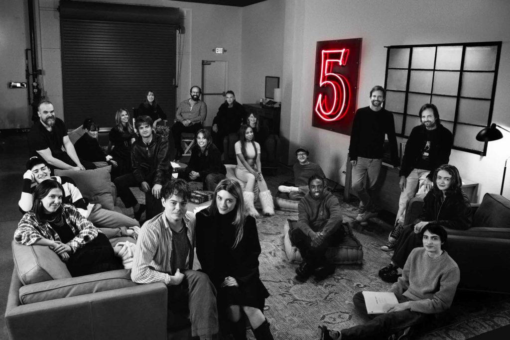 Black and white image of the Stranger Things cast on the set of season 5, with a neon red '5' sign in the background