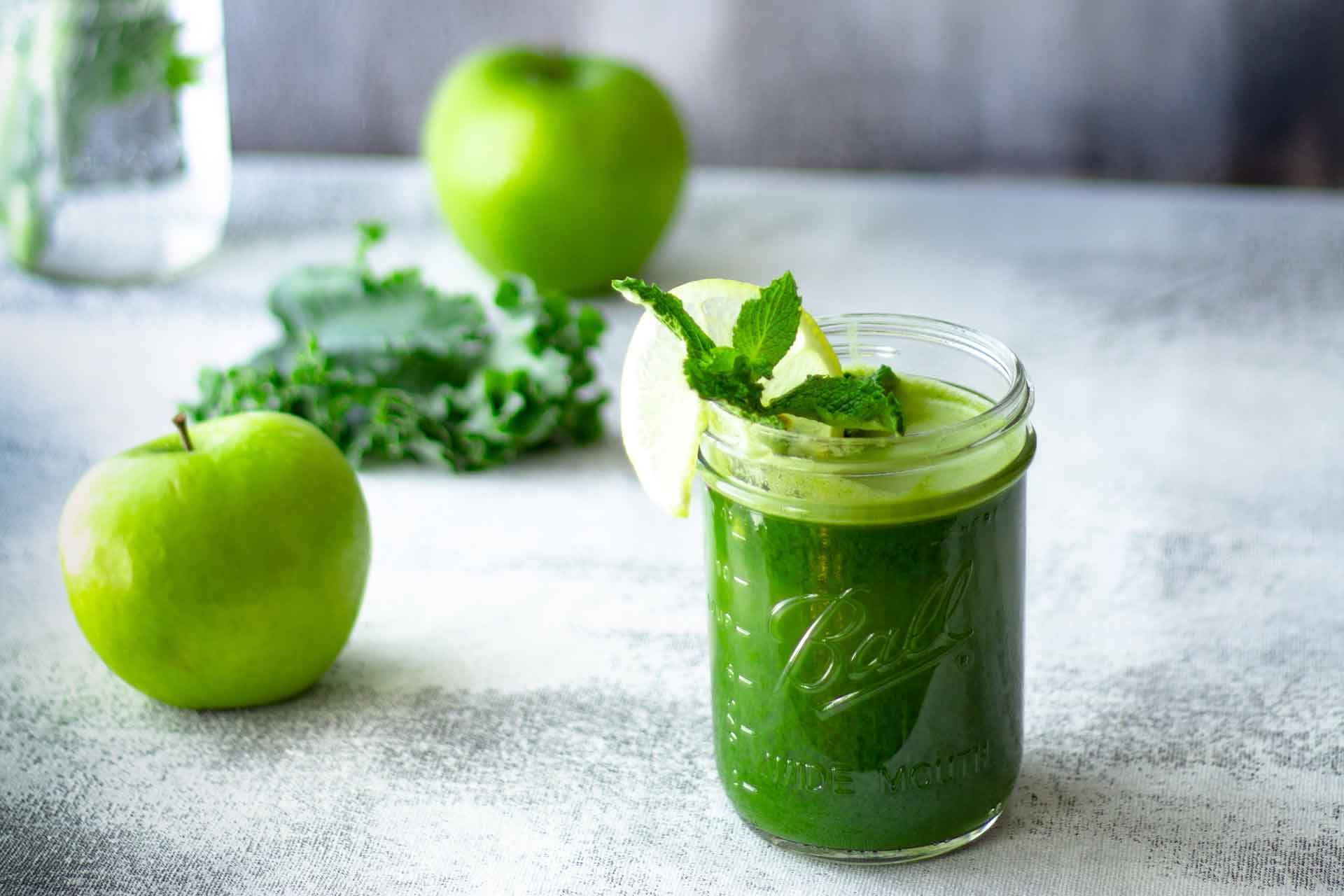 Mason jar of green juice with green apples and kale in the background.