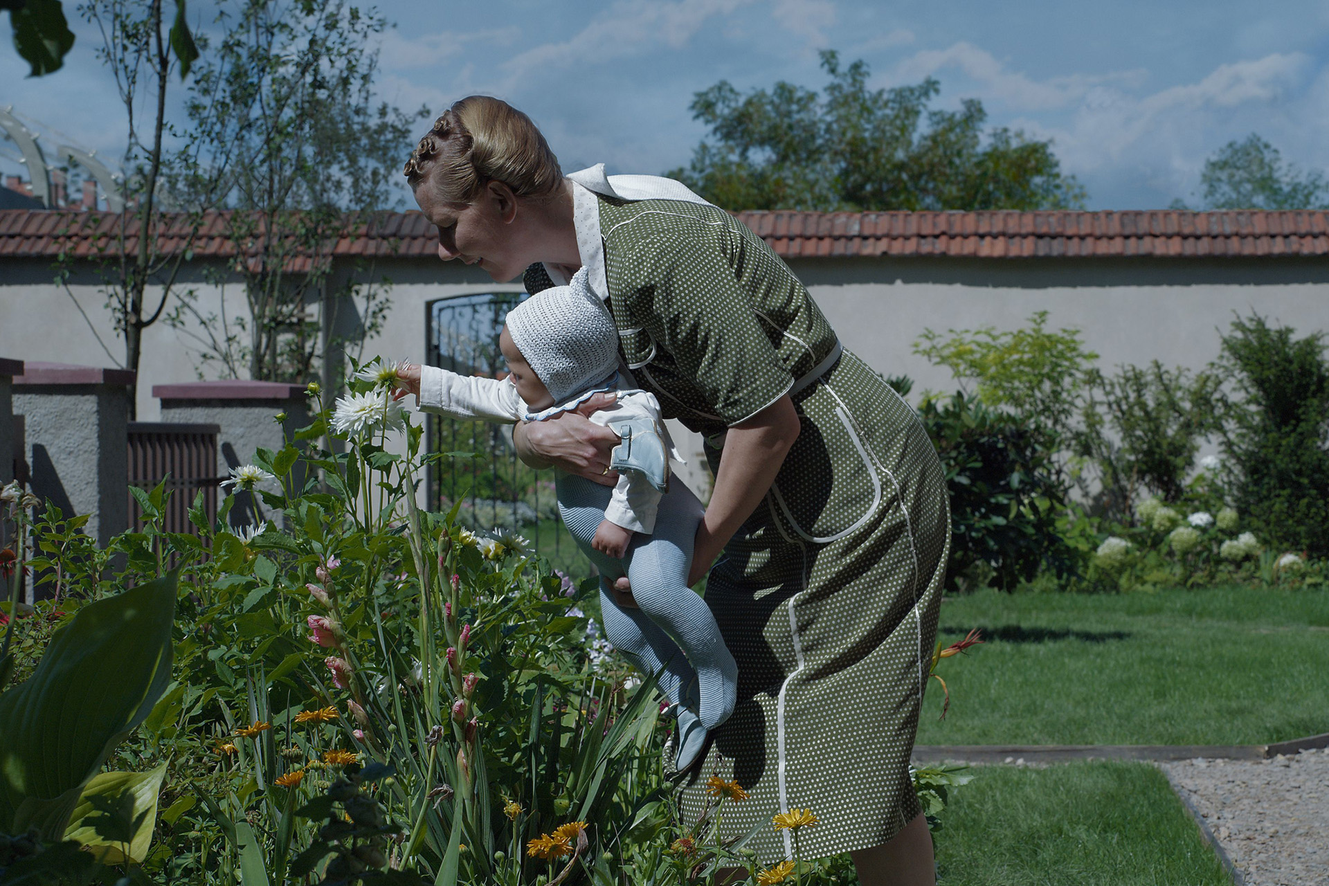 A still from The Zone of Interest depicting a woman holding a baby beside some plants